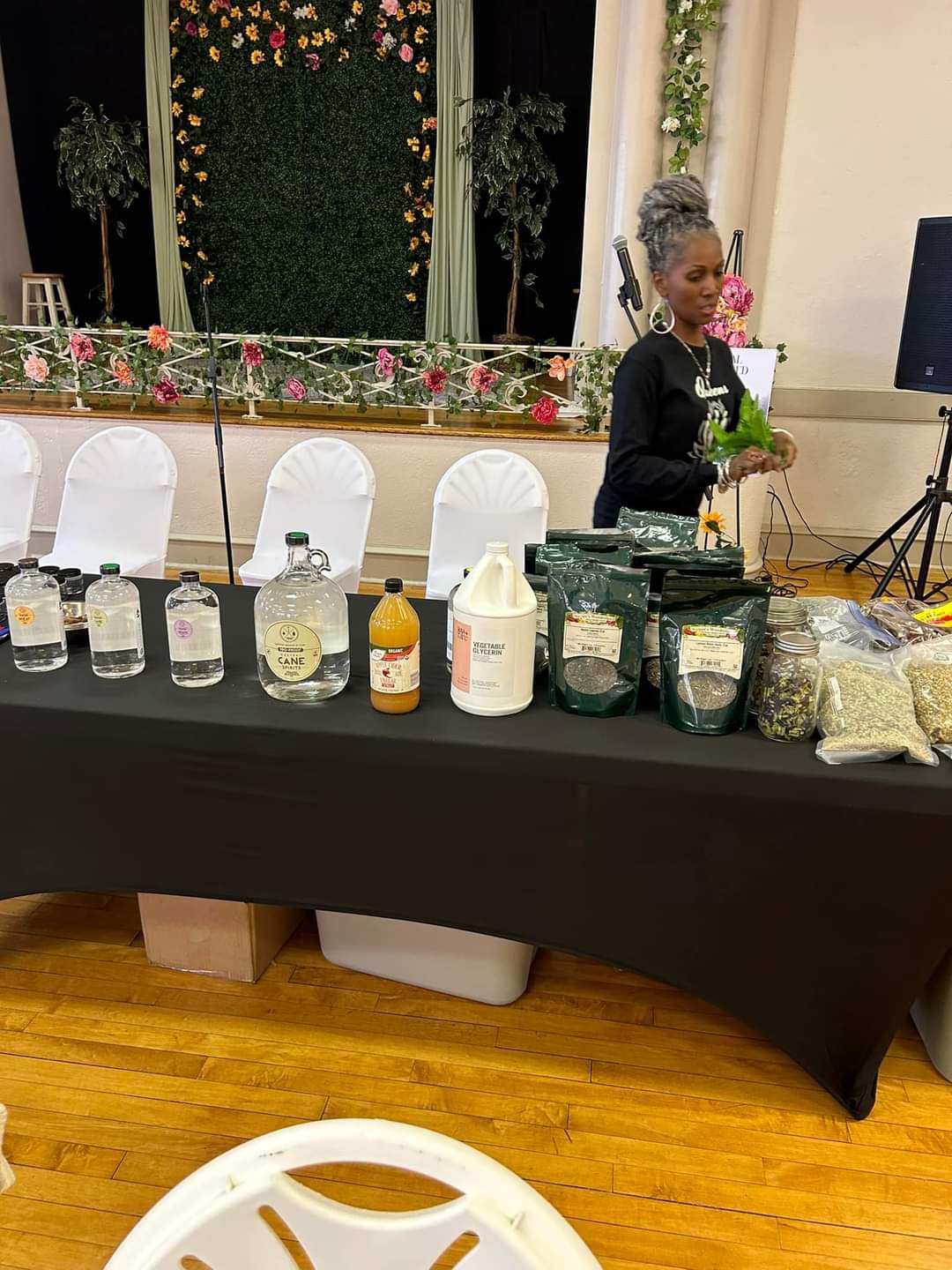Woman with gray hair and glasses stands behind a table with various jars and bottles, in a decorated indoor setting.