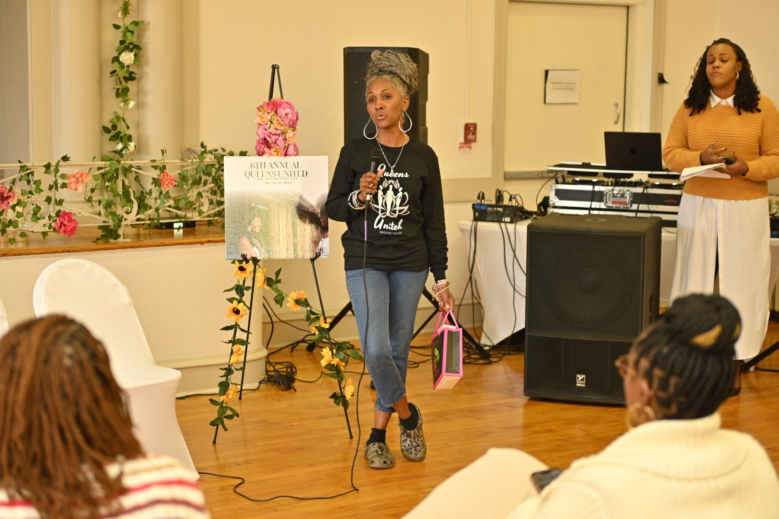 Woman speaking to an audience in a room with floral decorations and a sign on an easel, with another woman standing nearby.