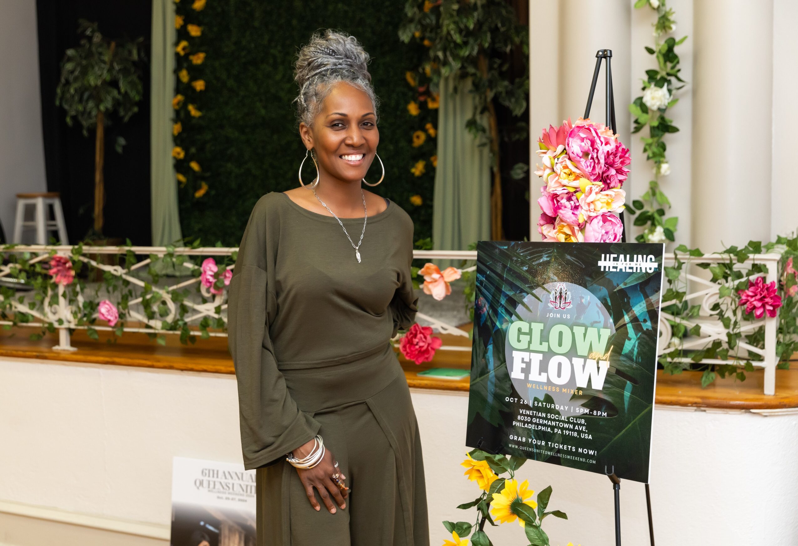 Smiling woman with gray hair in an olive green dress standing next to a colorful sign at an indoor event, decorated with flowers.