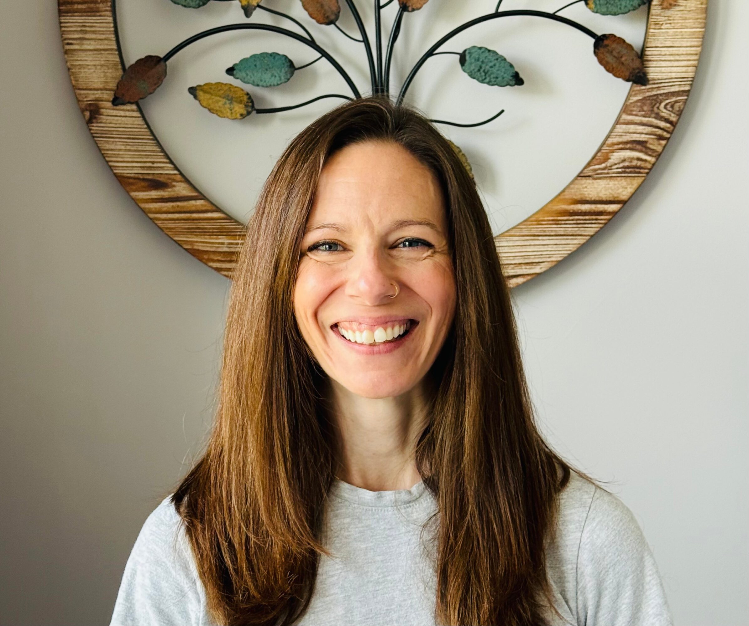 Smiling woman with long brown hair in front of a decorative wall hanging, wearing a light gray shirt.