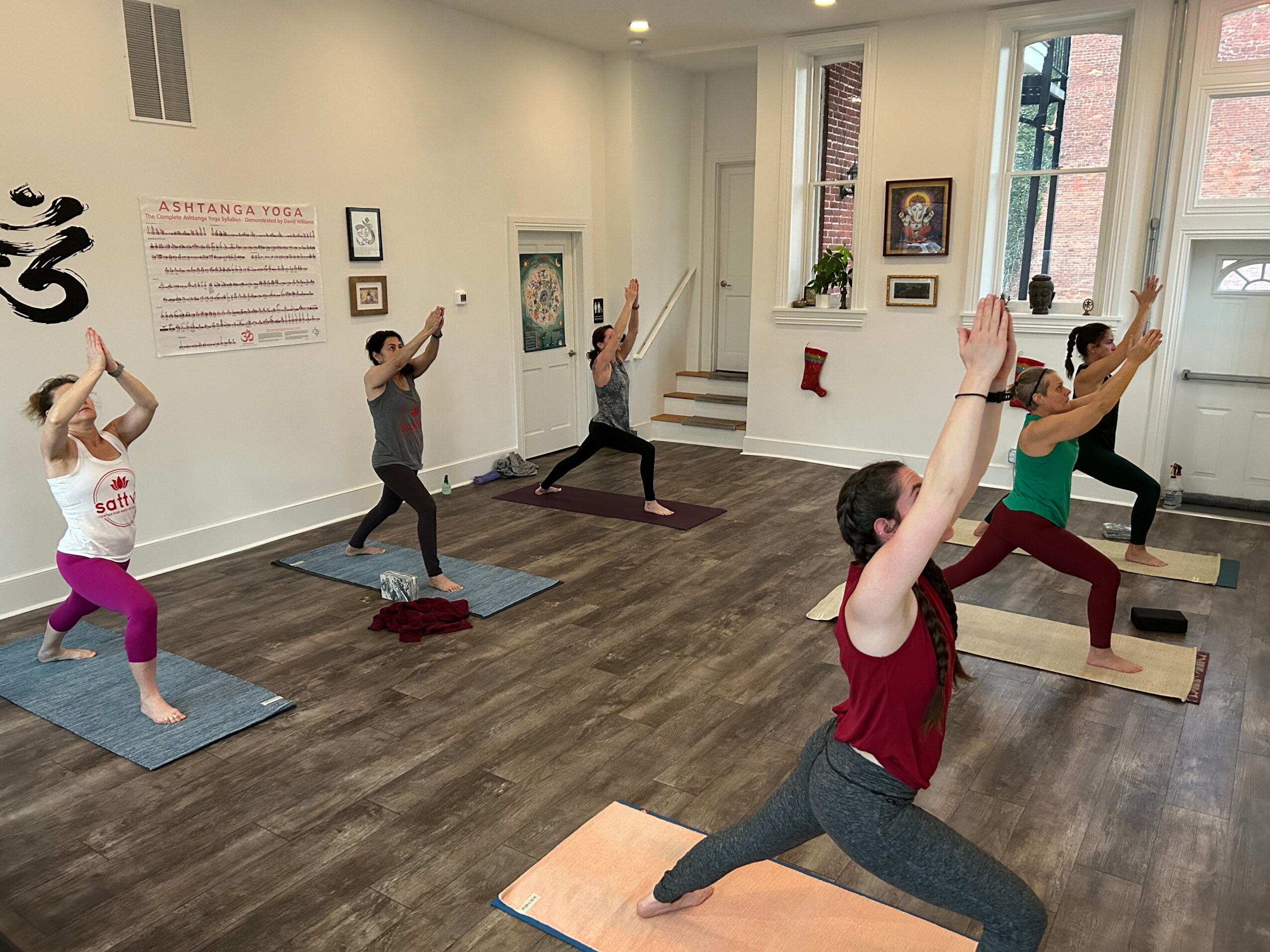 Group of people practicing yoga in a bright studio with large windows, mats, and a white wall with text and artwork.