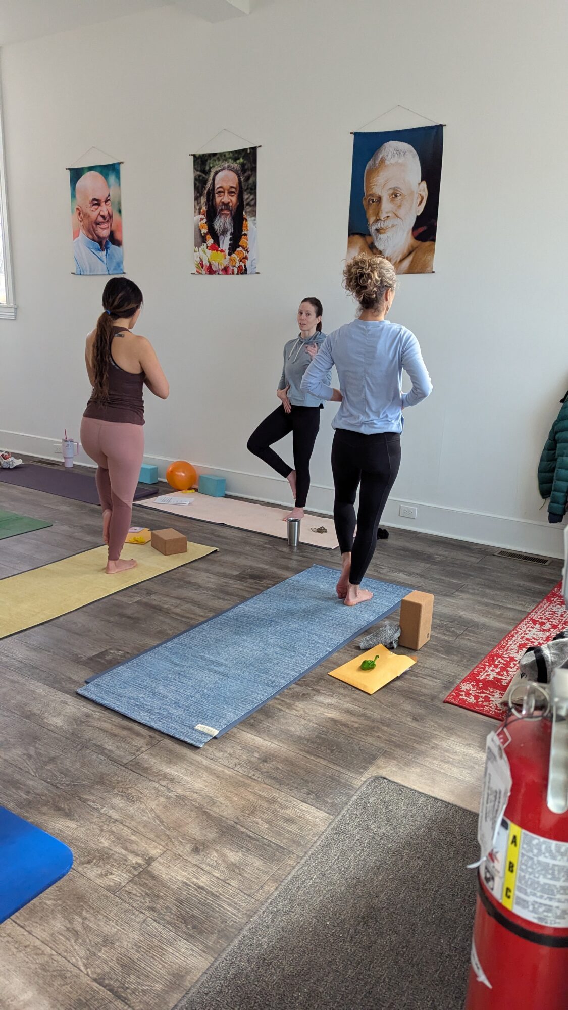 Three people in a room with yoga mats, posters on the wall, and a fire extinguisher nearby.