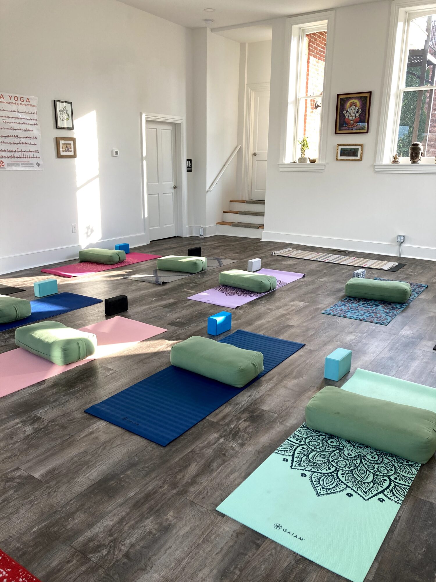 Yoga mats and cushions arranged on a wooden floor in a bright room with windows and artwork.