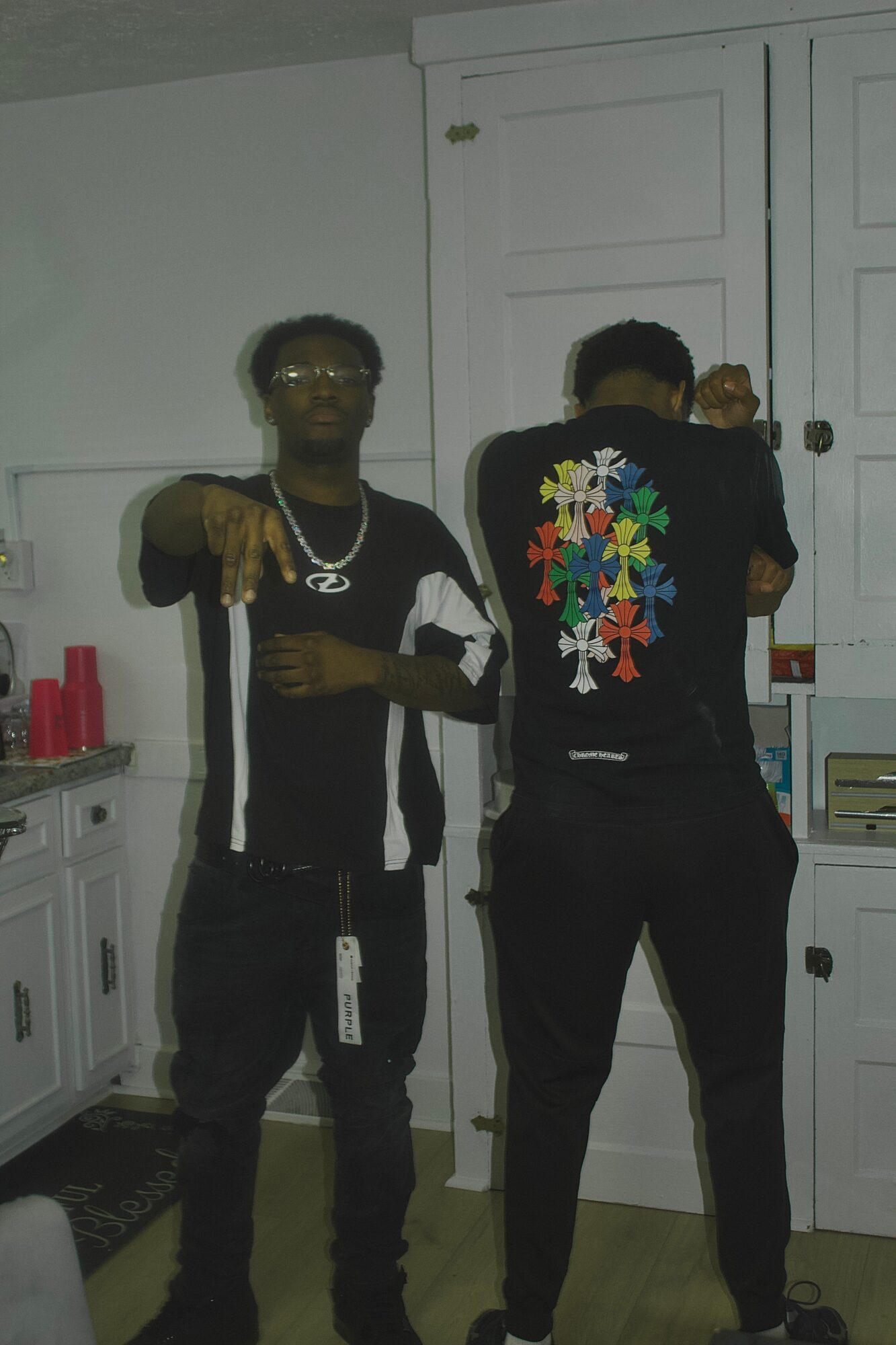 Two people taking a mirror selfie in a kitchen, one facing forward and the other with their back turned, both wearing dark clothing.