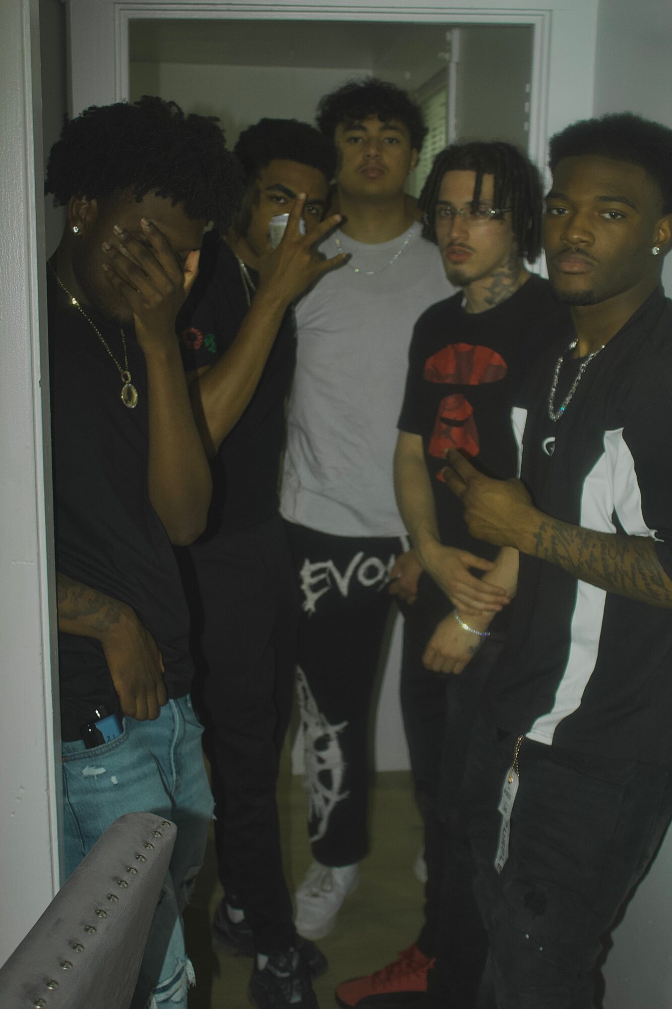 Group of five young men standing indoors, some with tattoos and jewelry, posing for a photo.
