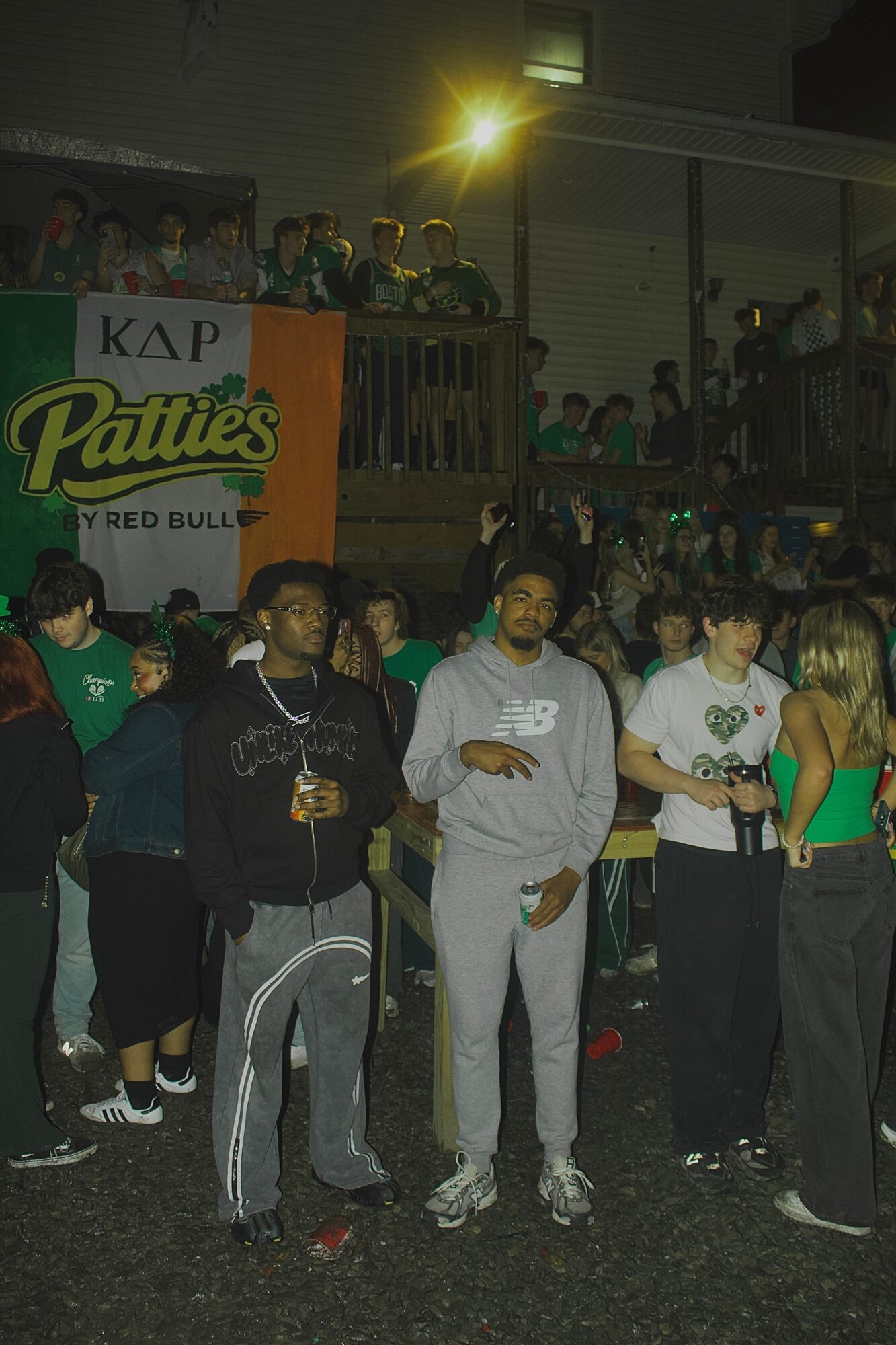 Group of people gathered outdoors at night, some holding drinks, with a building and bright light in the background.