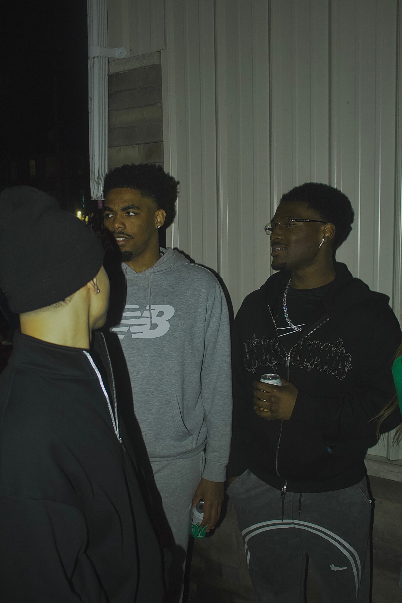 Three young men standing outdoors near a building with vertical siding, engaged in conversation, one holding a drink.