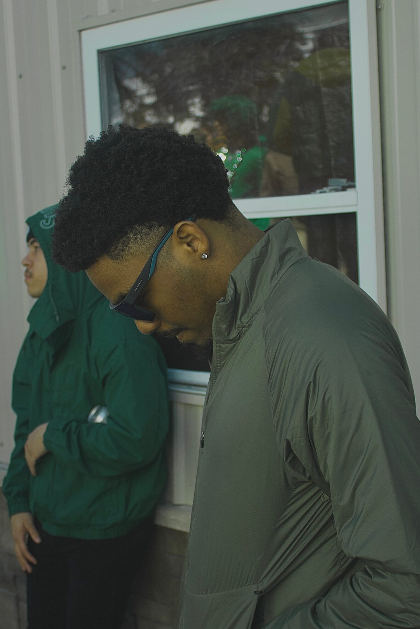 Two people standing near a window, one wearing glasses and a gray jacket, the other in a green hoodie, looking down.