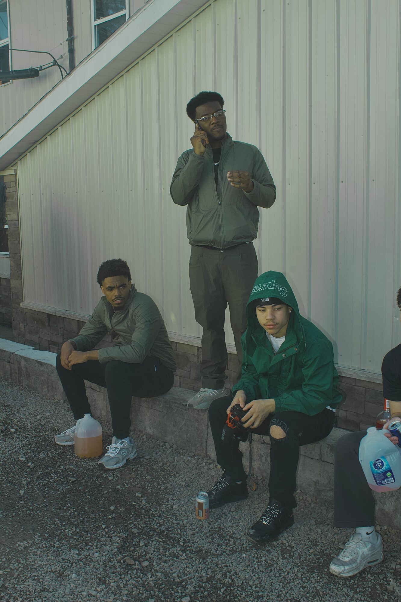 Three young men pose outdoors near a white building, one standing and two sitting, with casual clothing and accessories.