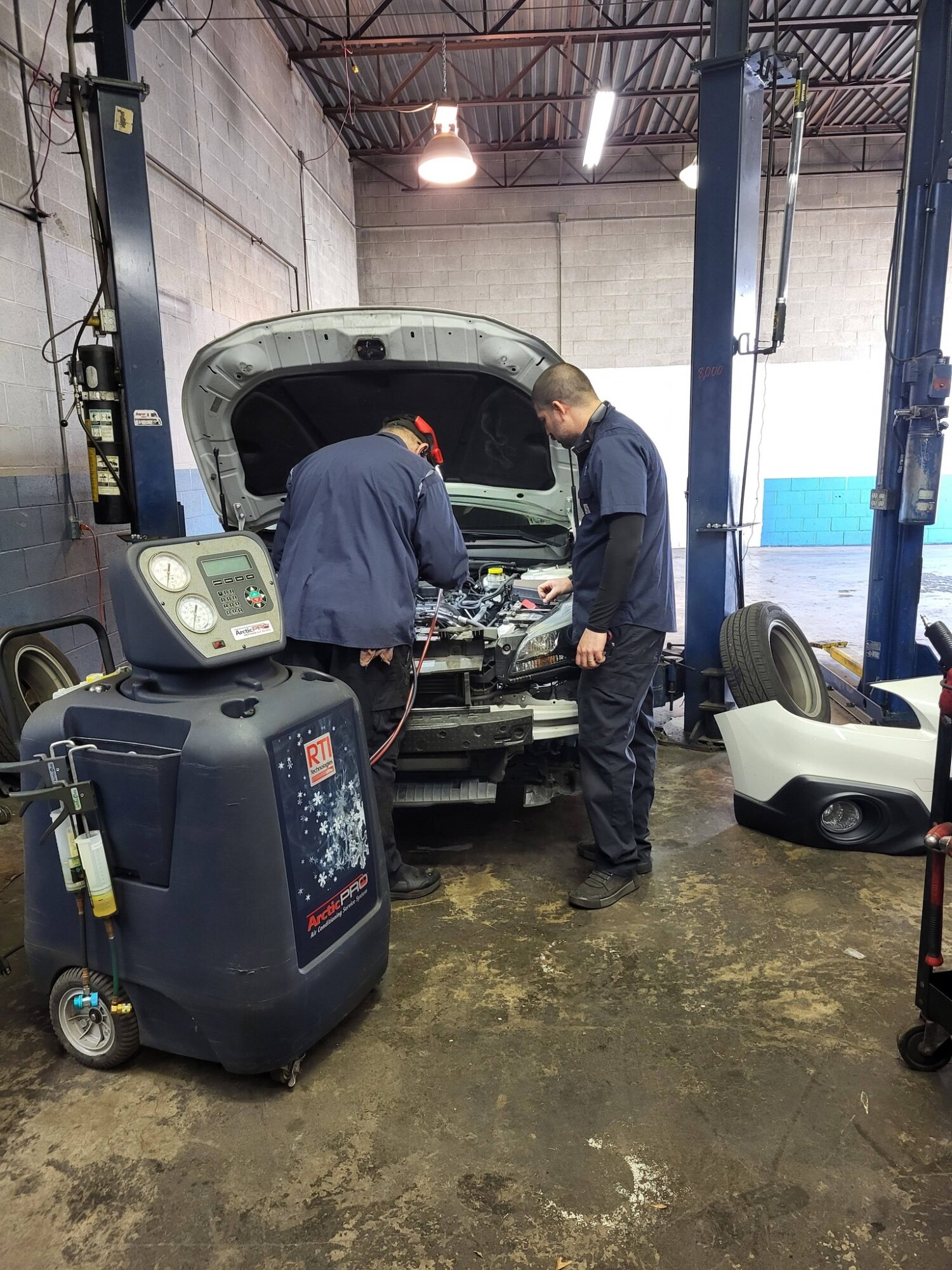 Two mechanics work on a car engine in a garage, with equipment and tools around them.