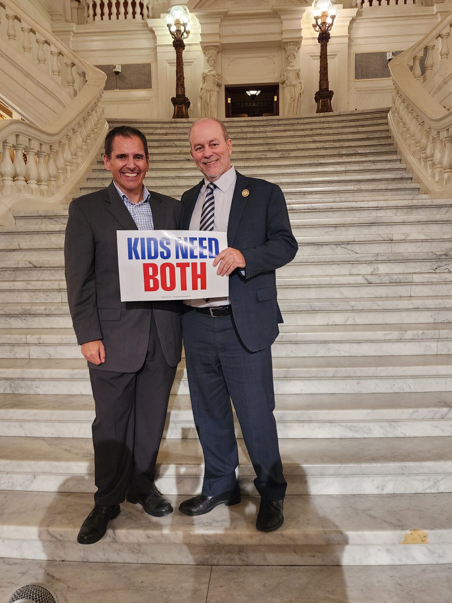 Two men in suits holding a sign that reads 'MUSLIM BOSTON' in a grand building with marble stairs.