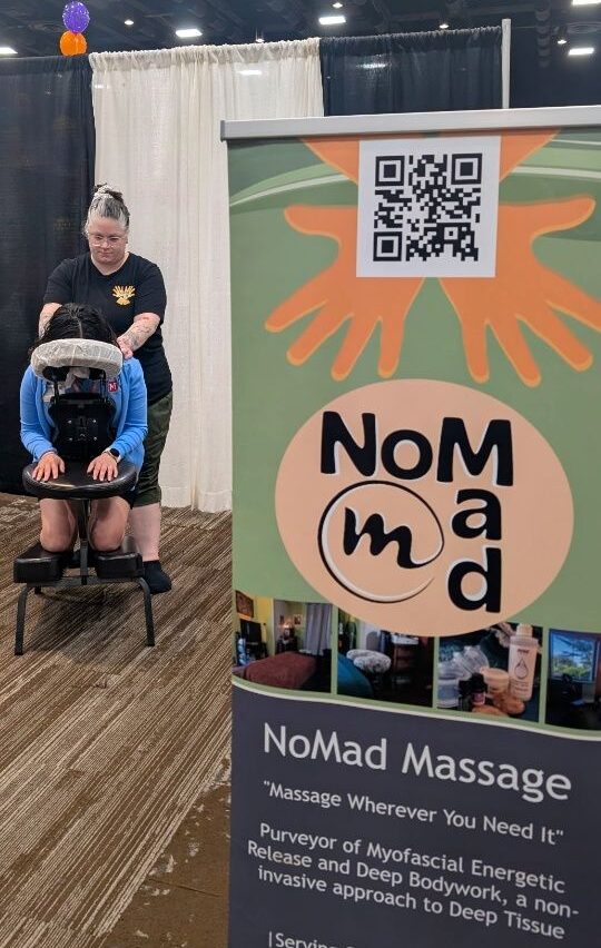A person receiving a massage from a therapist at a booth with a NoMad Massage sign, QR code, and colorful design.
