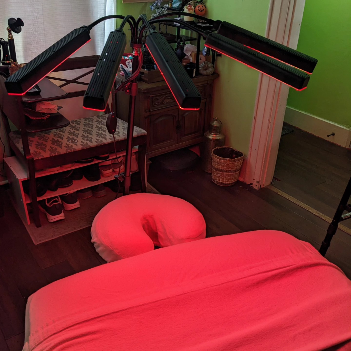Room with a bed, a massage table, and a shelf with shoes, illuminated by red light, with green walls and wooden floor.