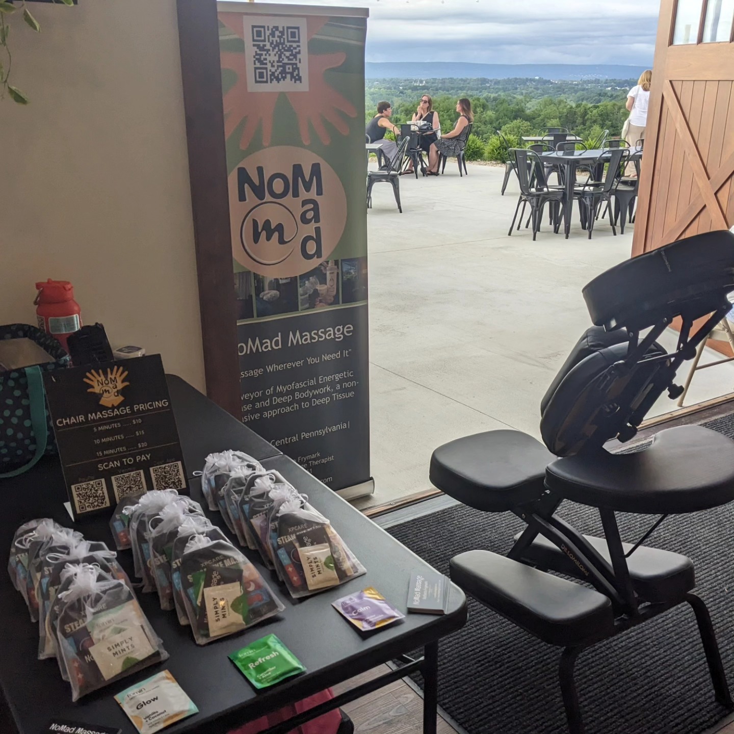 Indoor massage chair next to a table with massage products, open door leading to outdoor patio with tables and view, sunny day.