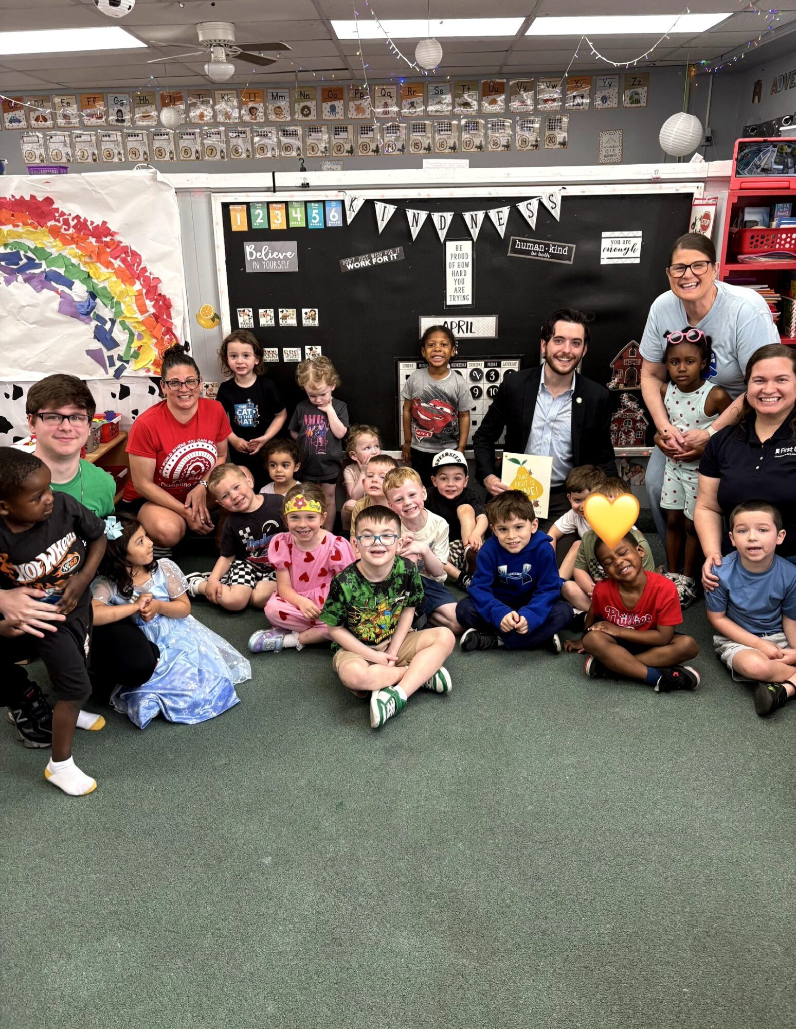 Group of children and adults in a classroom, some holding paper hearts, with a rainbow and blackboard in the background.