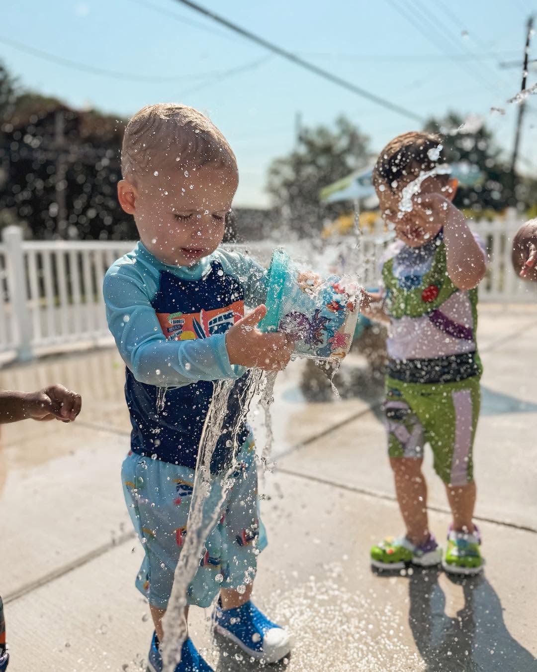 Two children playing with water outdoors, splashing water, with a white fence and trees in the background.