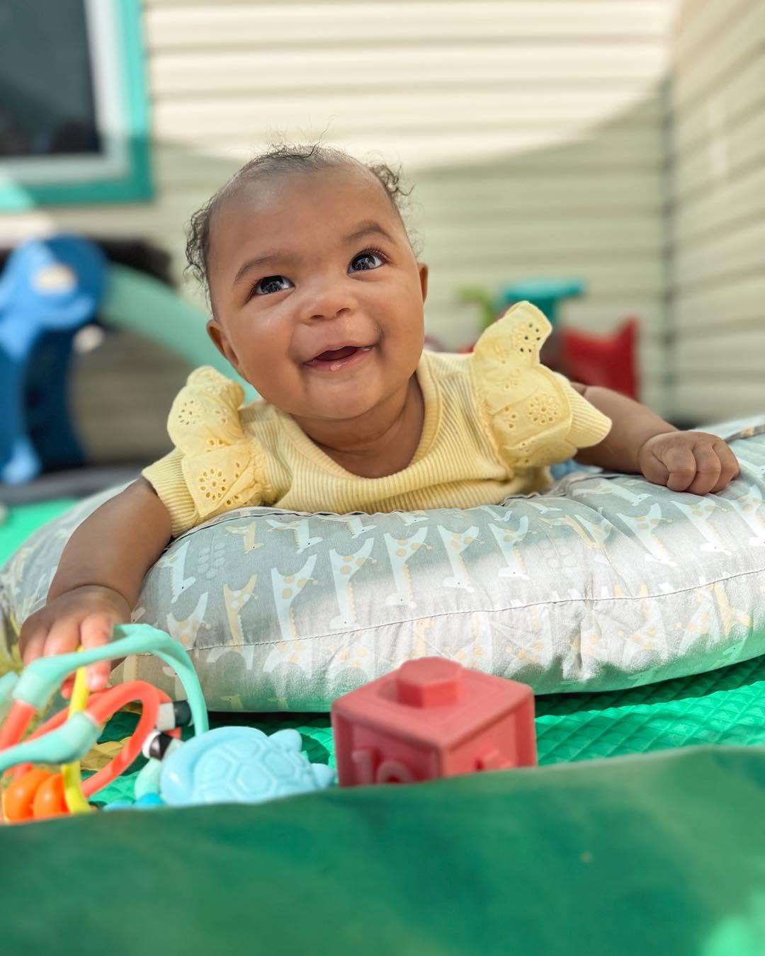 Smiling baby lying on a pillow outdoors, wearing a yellow dress, with toys nearby, in a backyard setting.