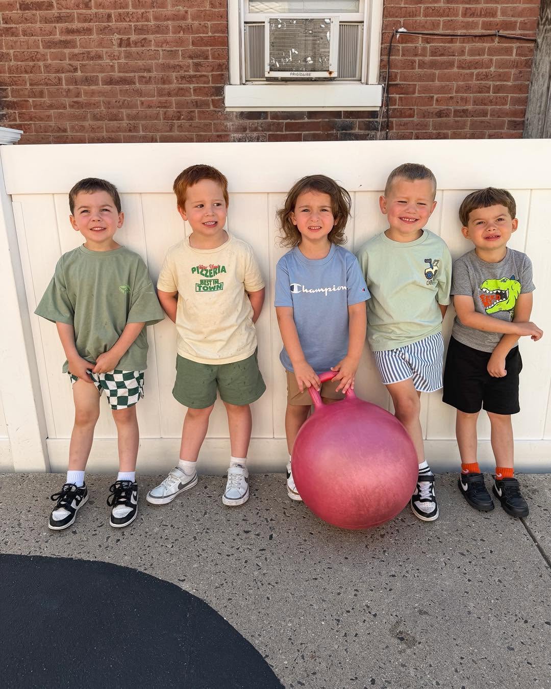 Five children stand in front of a white fence, smiling, with a brick building and window behind them, one holding a pink ball.