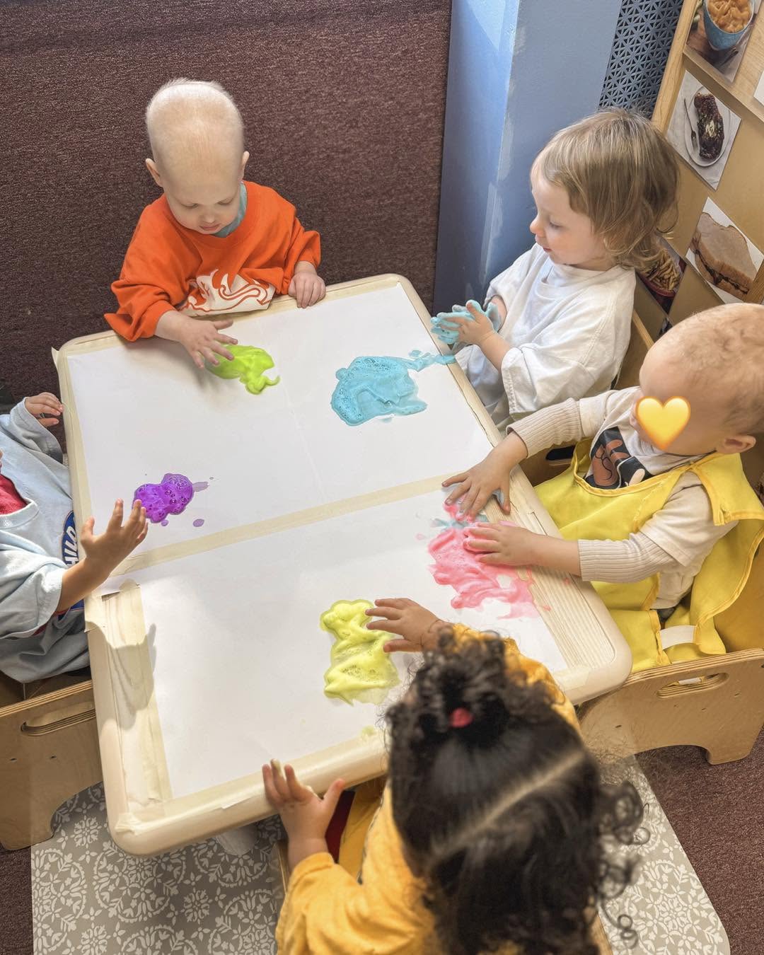 Four children sit around a table with colorful slime, engaging in a sensory activity, with a dog nearby. One child has a yellow shirt, another a white shirt, and a third a red shirt. A girl with dark hair and a red bow is also present.