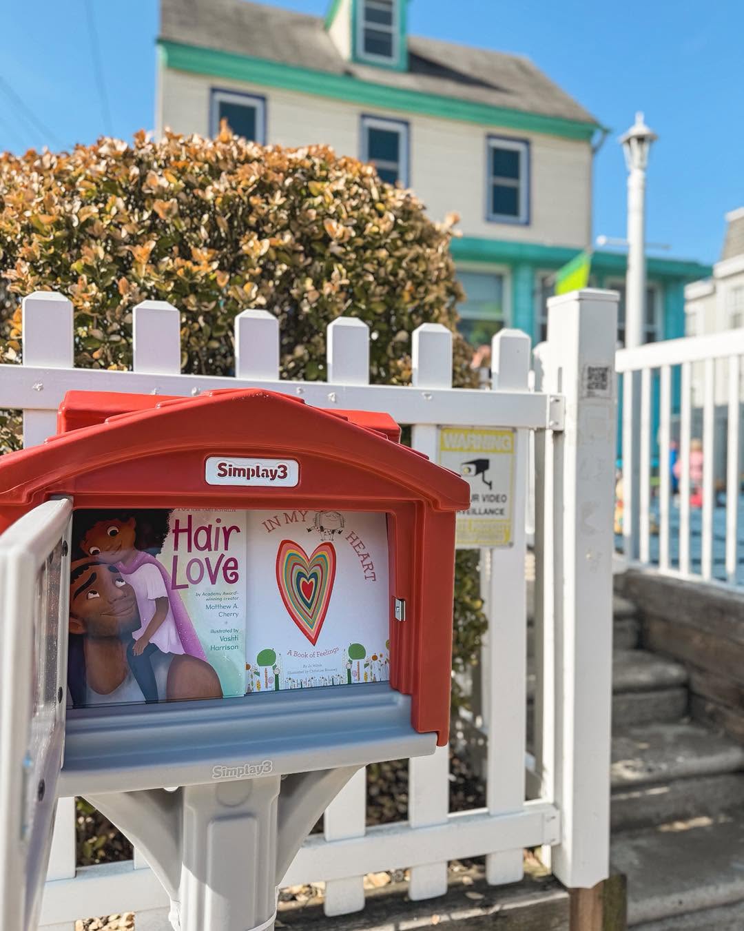 Red donation box with a colorful heart design and text, placed in front of a white picket fence and a house.