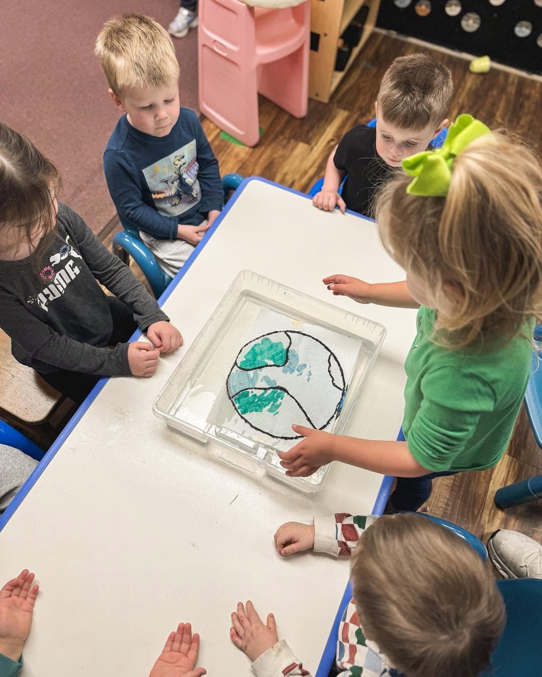 Children gather around a table with a clear container holding a drawing of a fish and water, engaging in an activity.