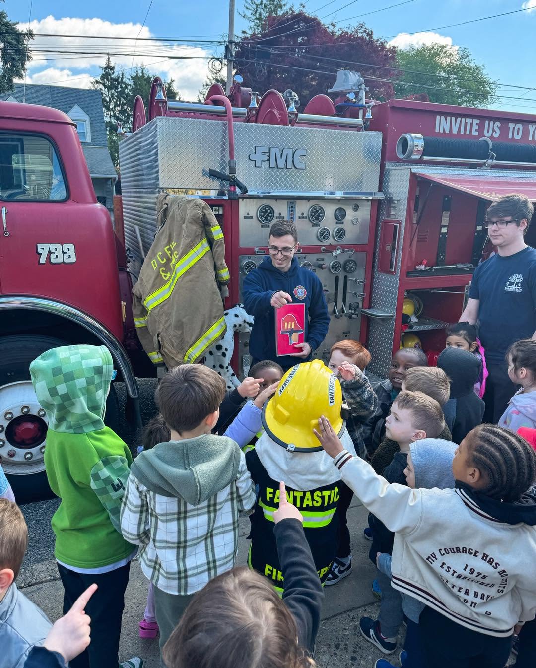 Firefighter shows a fire extinguisher to children gathered around a fire truck outdoors, with trees and sky in background.