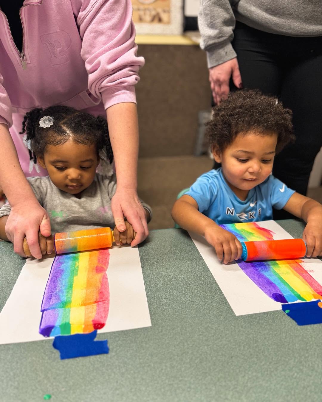 Two children create rainbow art with colorful paper strips on white paper, guided by adults. The children are focused on their artwork.