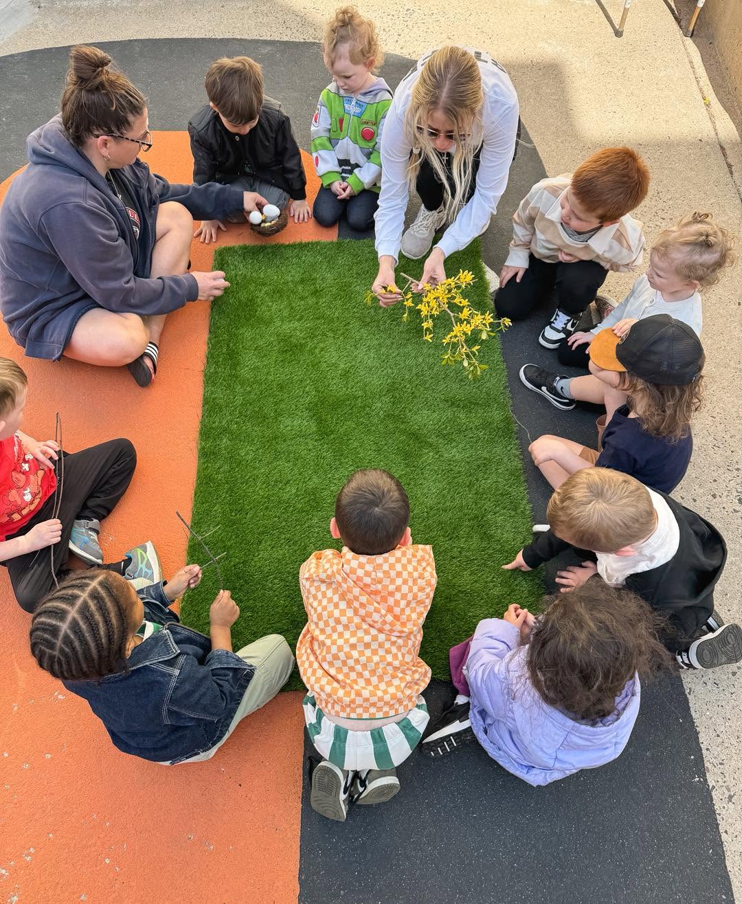 Group of children and an adult sitting in a circle around a small patch of artificial grass outdoors, engaging in an activity.