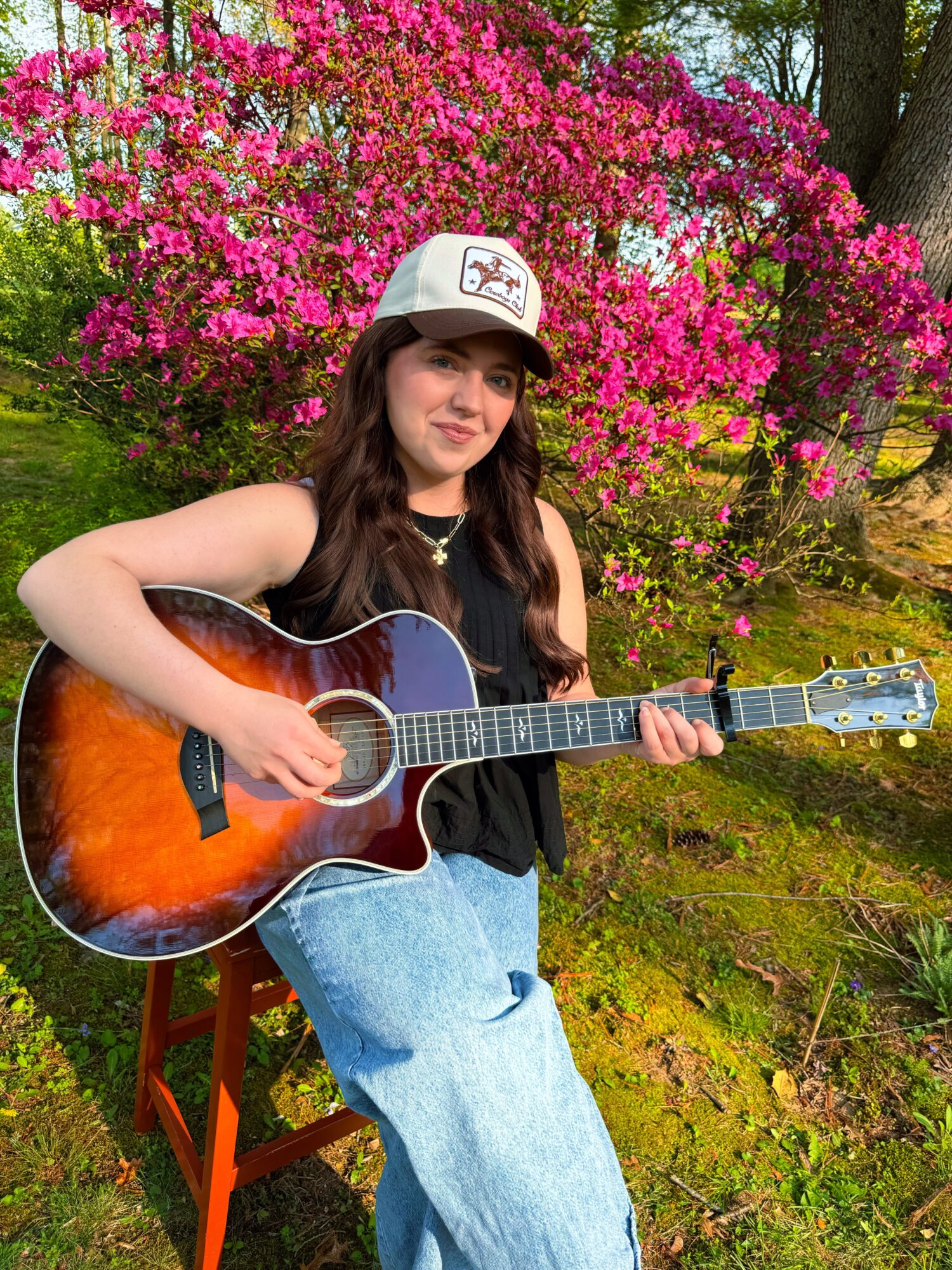 Young woman with long dark hair playing an acoustic guitar outdoors, pink flowering bush in background, wearing a white cap and black top.