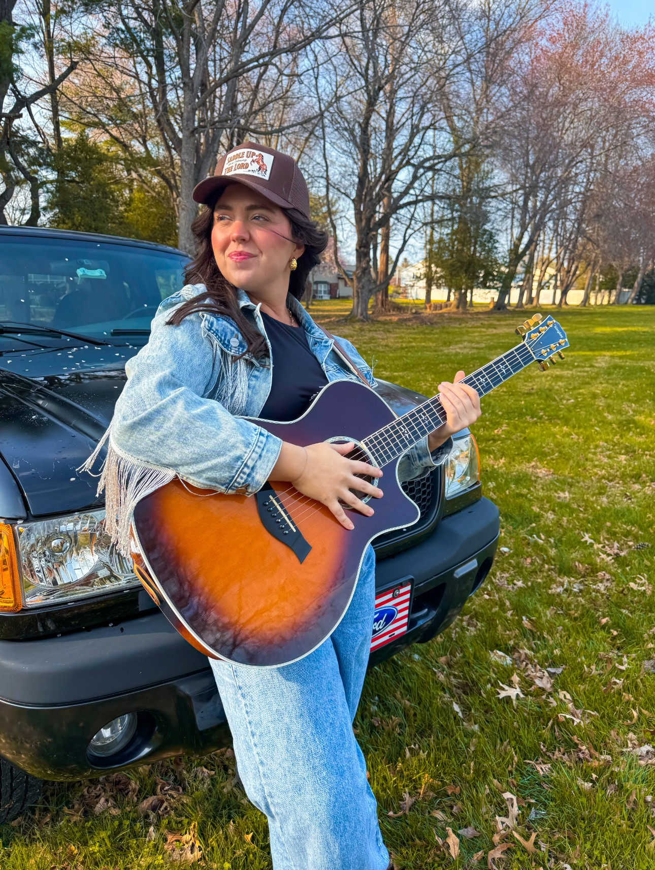Woman with curly hair wearing a cap, denim jacket, and jeans playing an acoustic guitar outdoors near a black vehicle, trees, and grass.