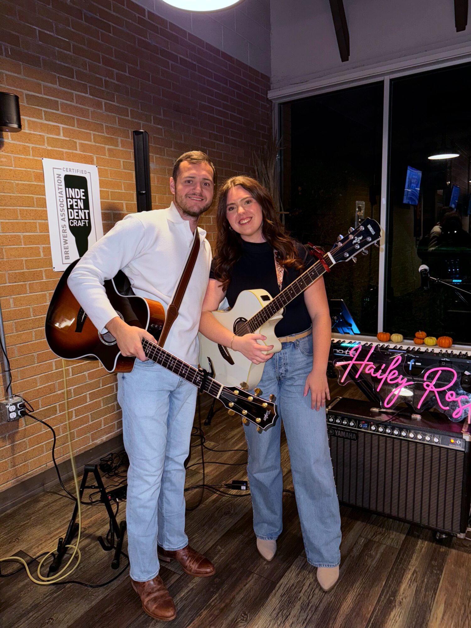 Two people holding guitars, standing indoors near a brick wall and a neon sign, smiling at the camera.