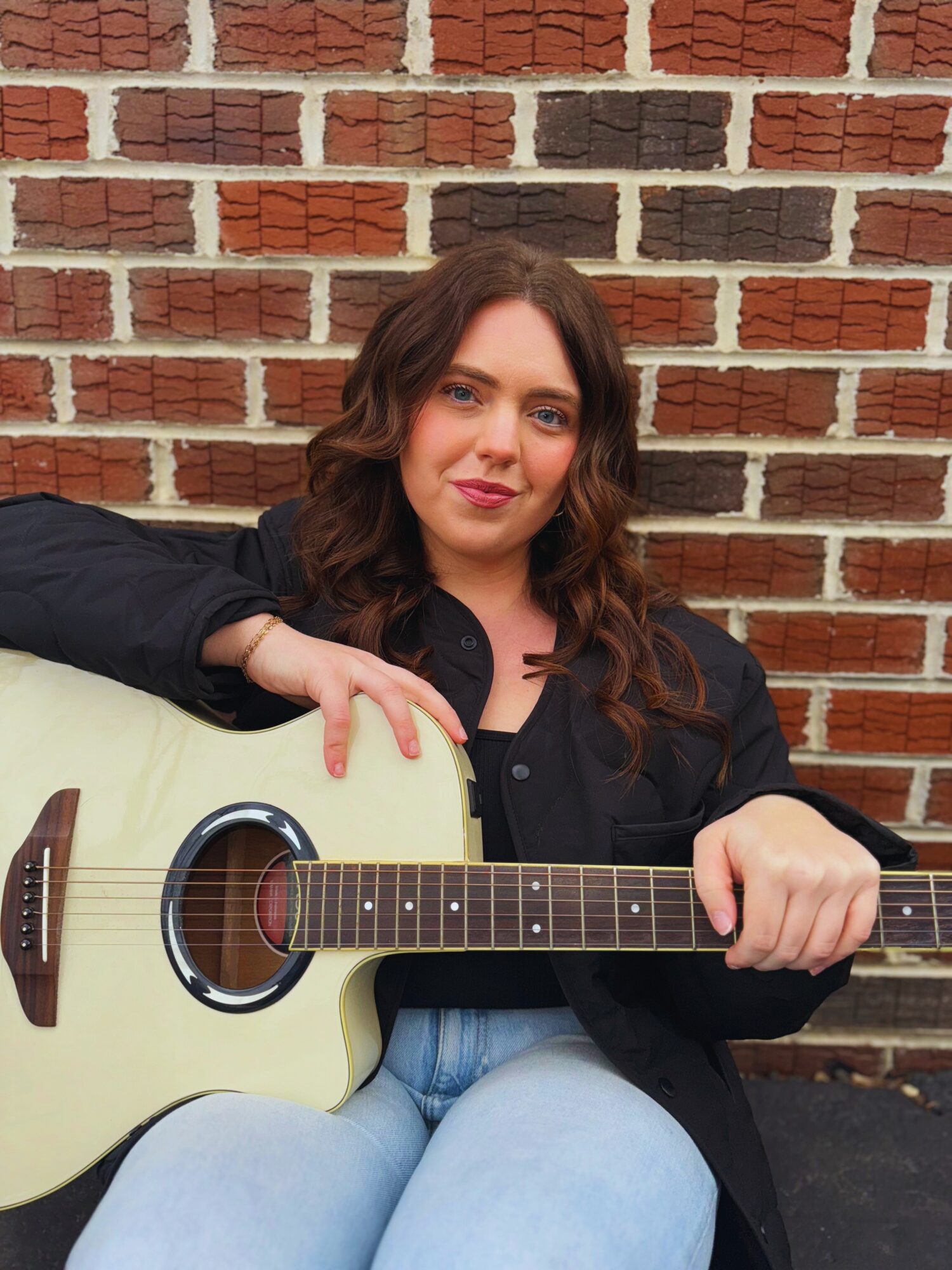 Woman with wavy brown hair holding a light-colored acoustic guitar against a brick wall.