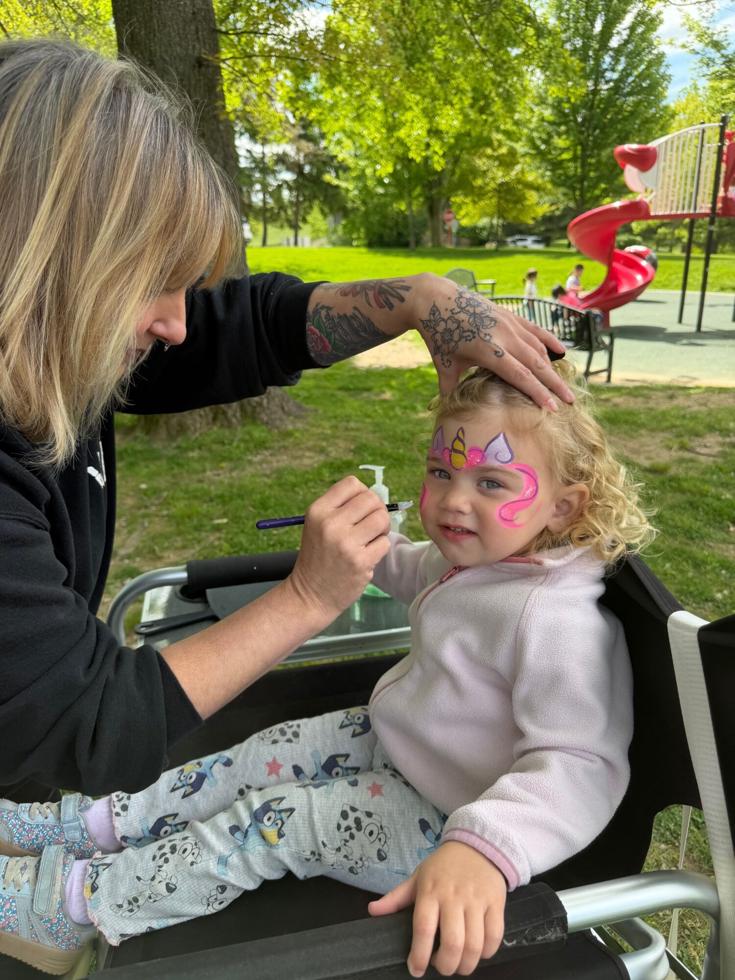 Child with face paint and curly hair smiling, leaning on a table outdoors, with a person with blonde hair and tattoos touching their face.