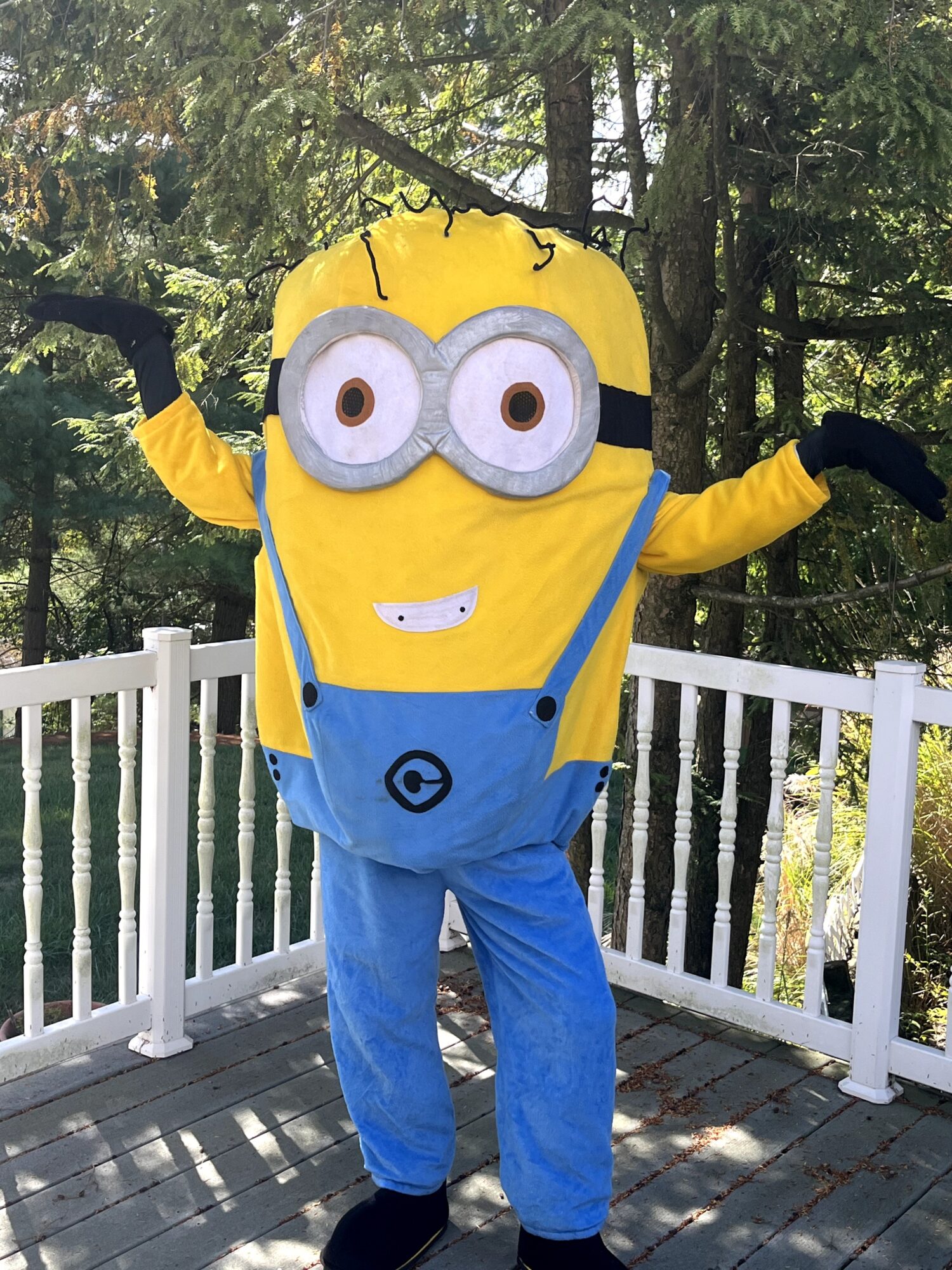 Person in Minion costume with arms raised, standing outdoors on a deck with trees in background.