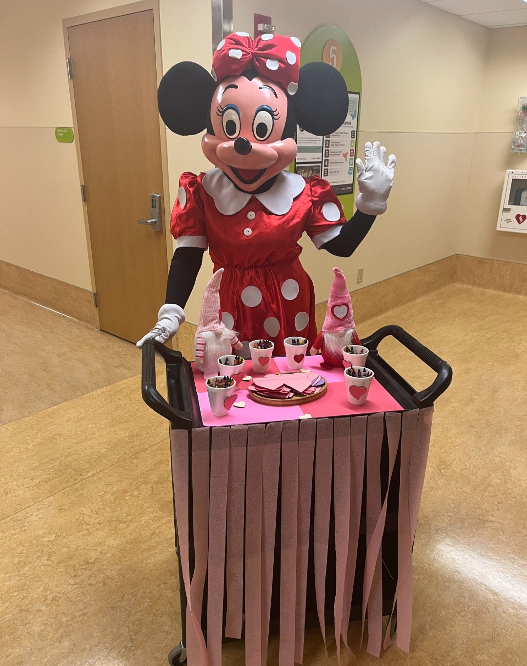 Minnie Mouse character in a red polka dot dress and bow, standing behind a pink table with tea set, waving.