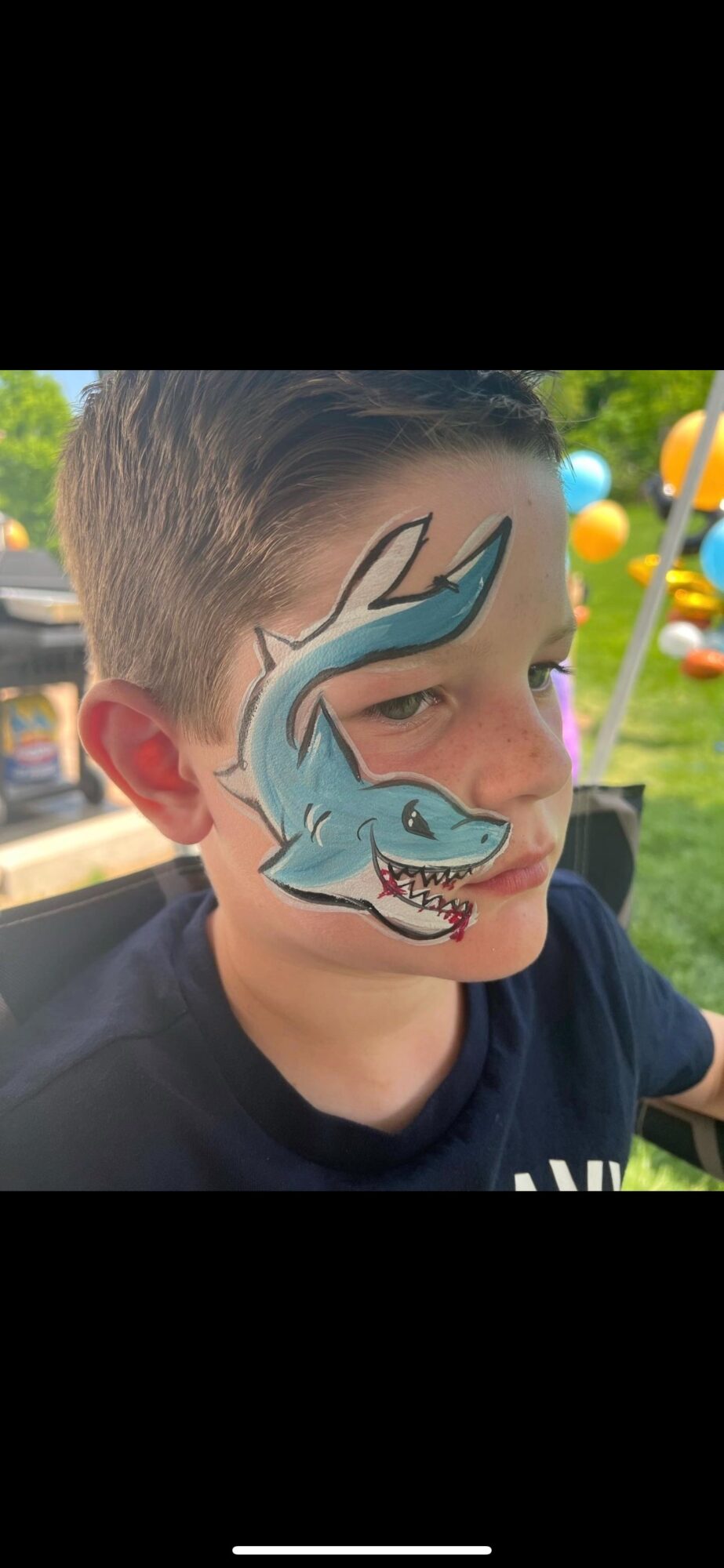 Child with face paint of a blue dragon, outdoors with colorful balloons in the background.