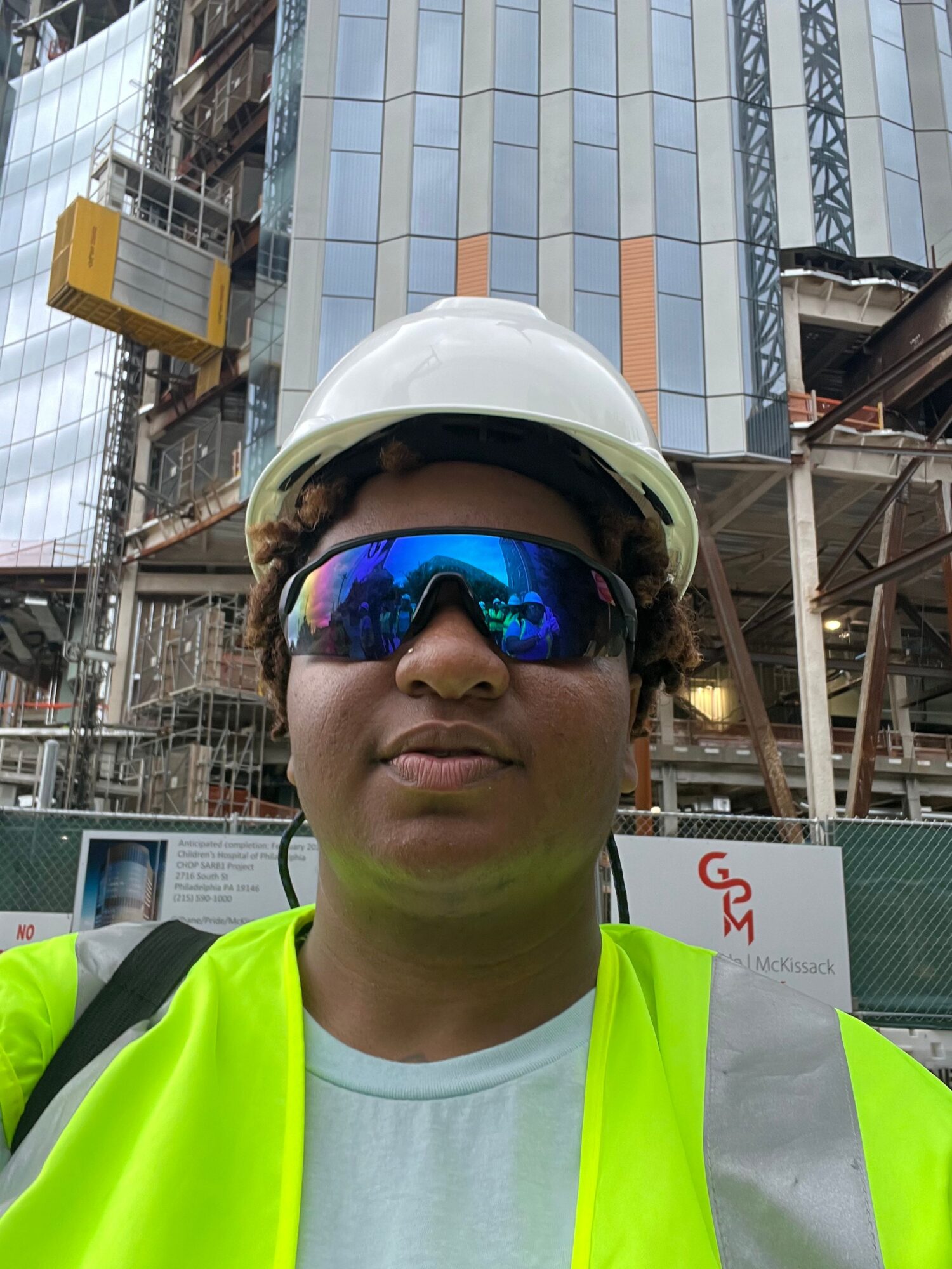 Person wearing a white safety helmet and reflective vest at construction site, with buildings and scaffolding in background.
