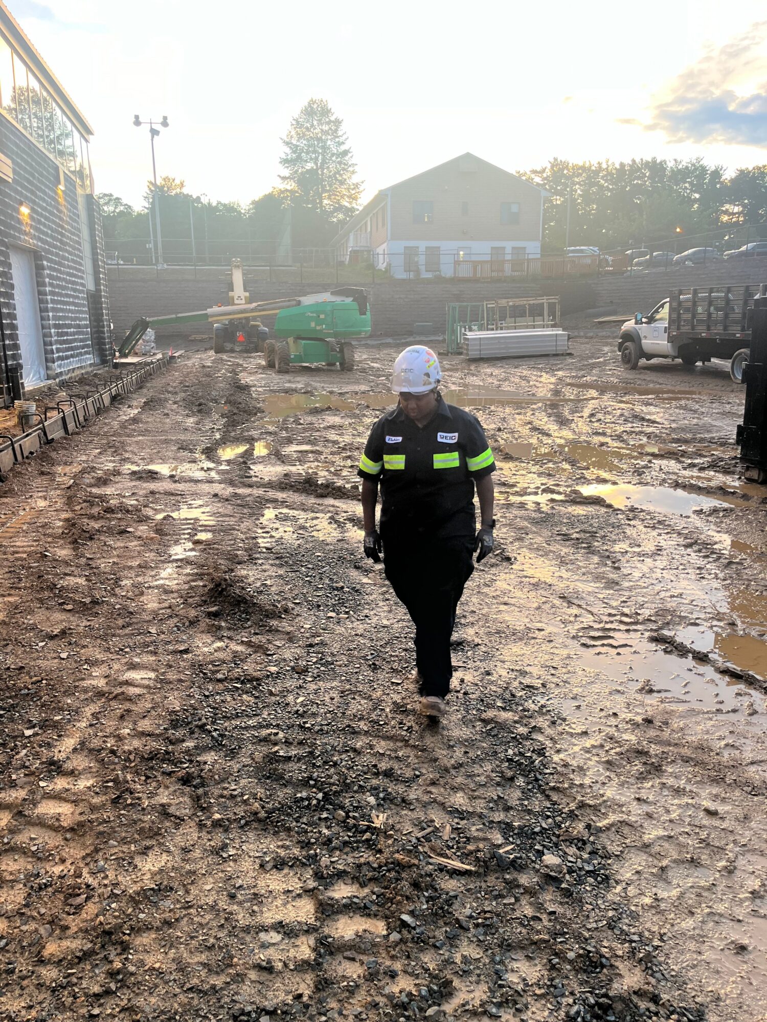 Person walking on a dirt road with buildings and trees in the background, wearing dark clothing and a cap.