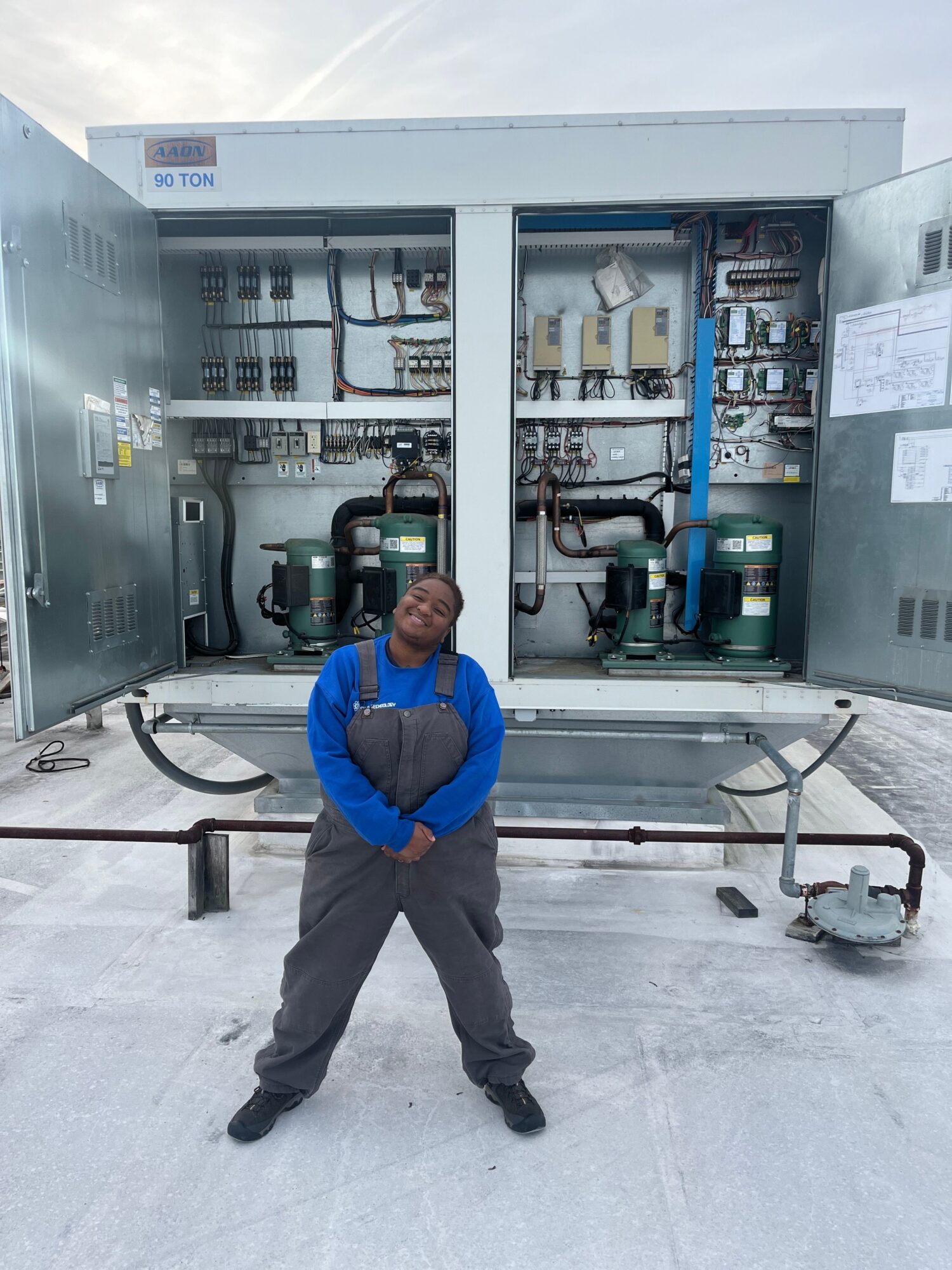 Man in blue shirt and gray pants standing in front of electrical equipment, holding a cart, in an industrial setting.
