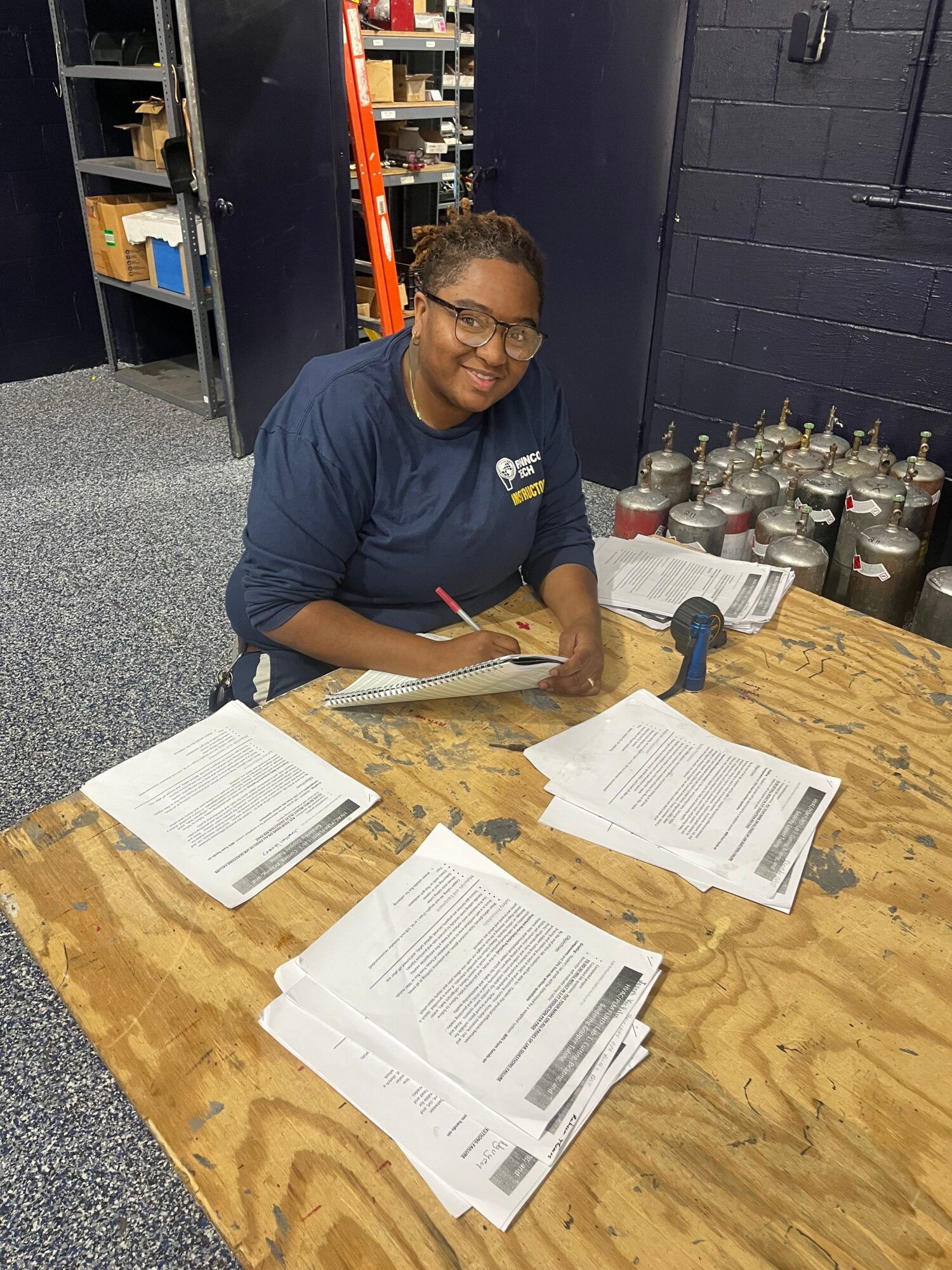 Person with glasses and dark hair, wearing a navy shirt, sitting at a wooden table with papers and documents, in a room with shelves and bottles.