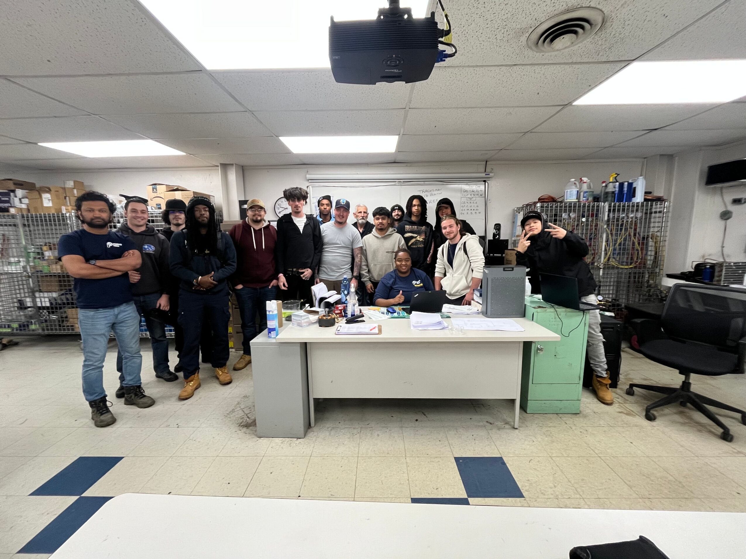 Group of people gathered around a table in a room with shelves and equipment, some standing and some sitting.