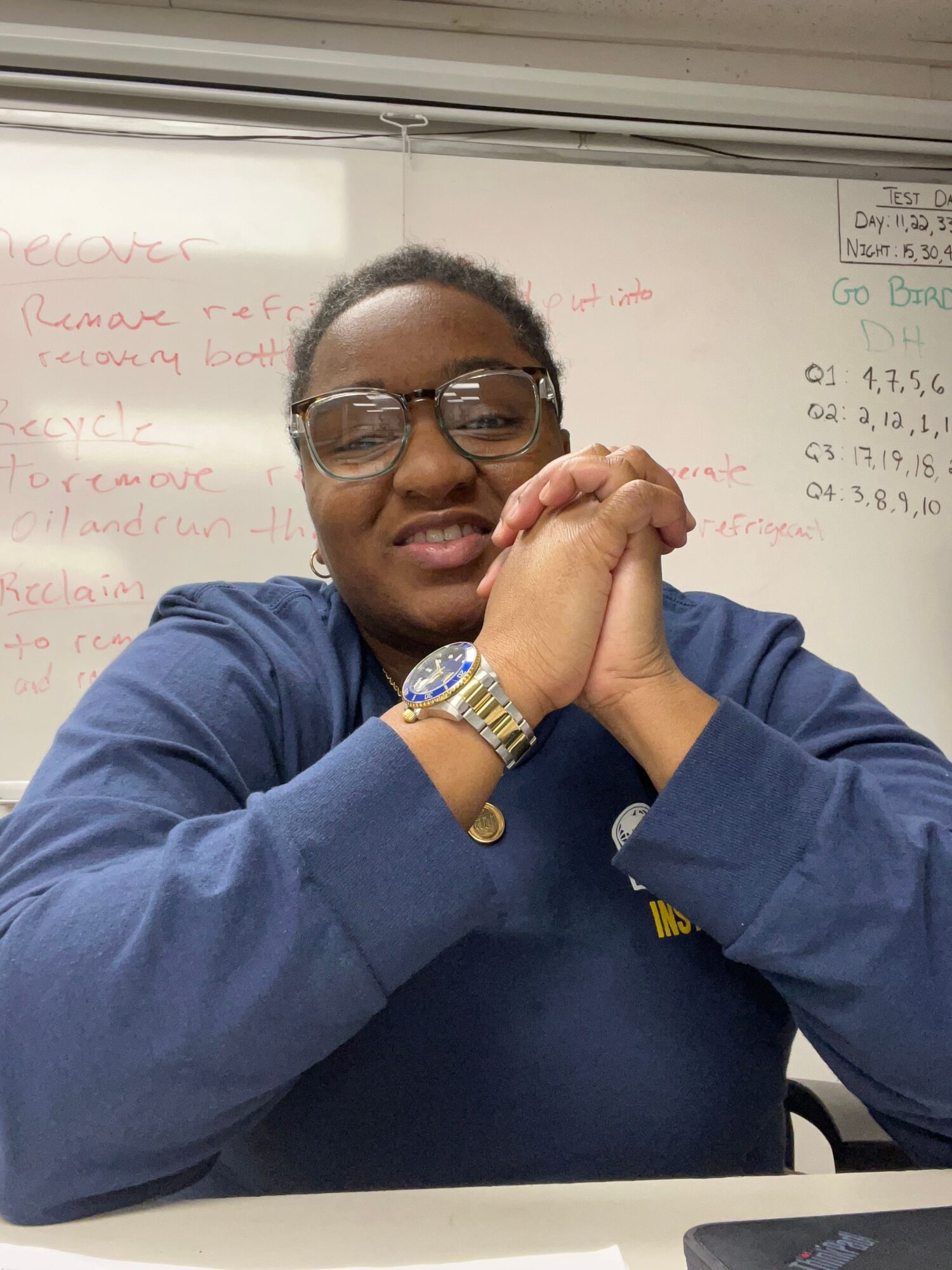 Person wearing glasses and a blue jacket rests their head on clasped hands on a desk, whiteboard with writing in background.