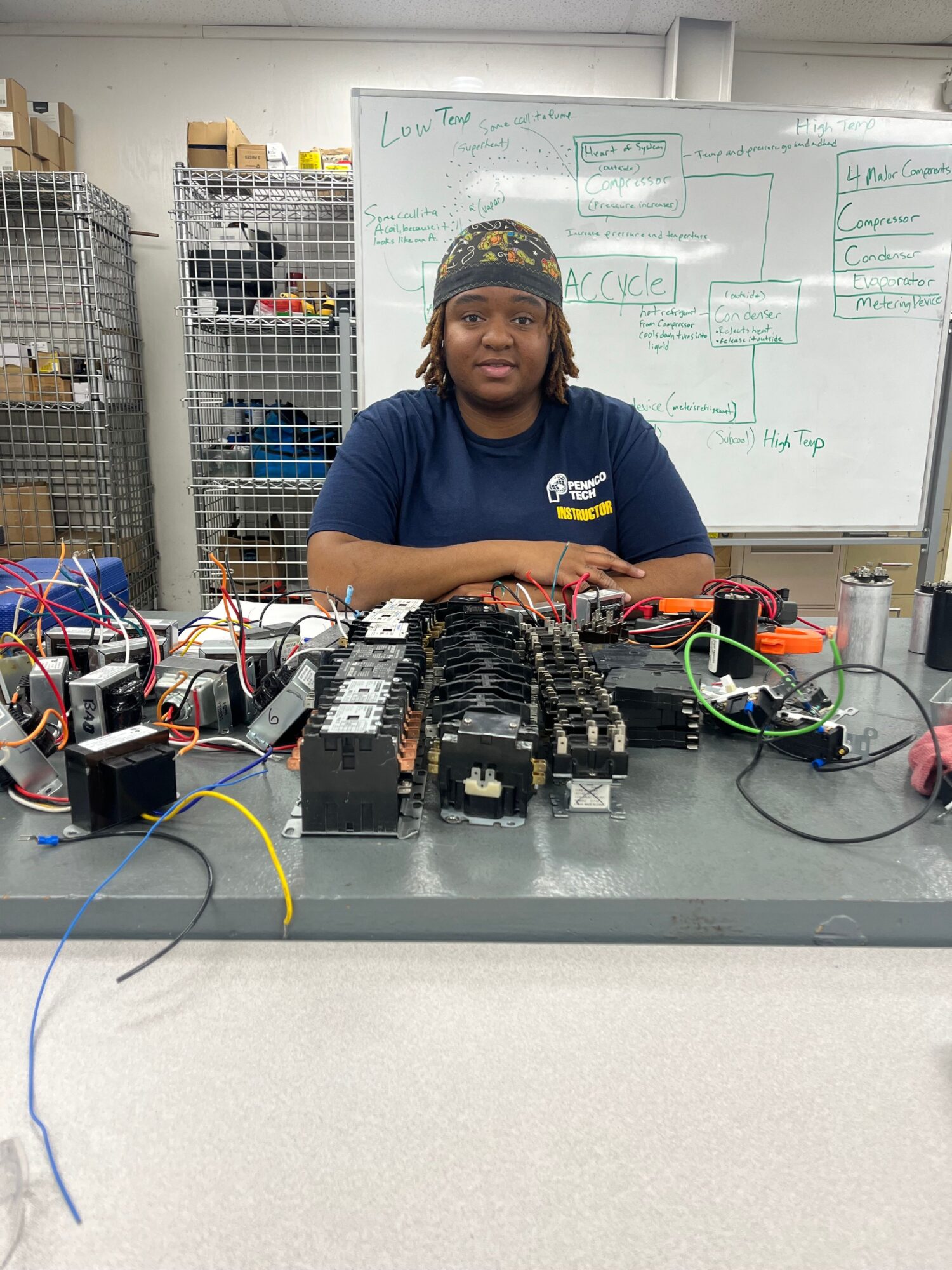 Person working with electronic components and wires on a table, whiteboard with diagrams in background, person smiling, wearing a cap and blue shirt.