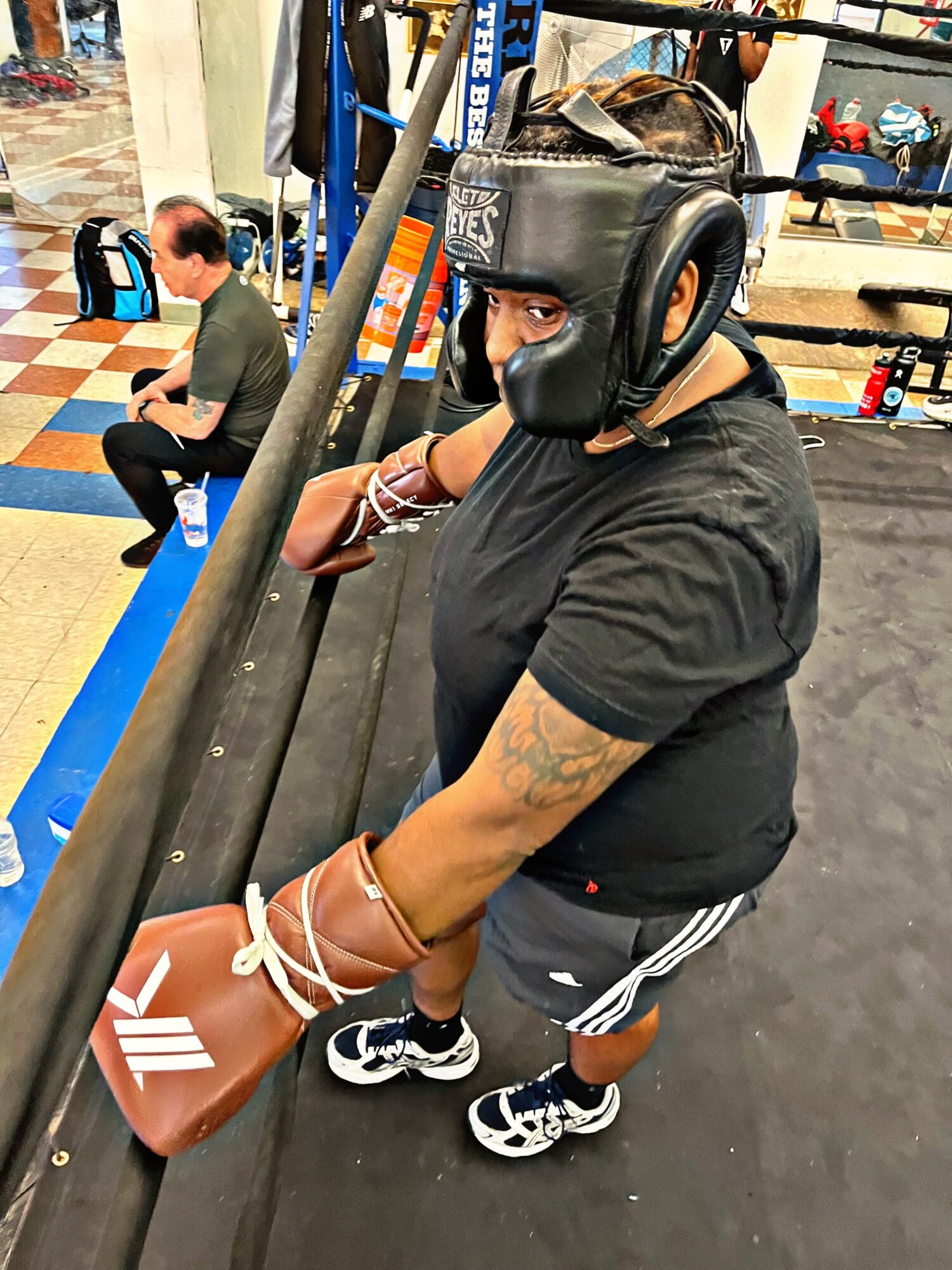 Person wearing a boxing helmet and black t-shirt, sitting on a bench in a gym, holding boxing gloves, with gym equipment in background.