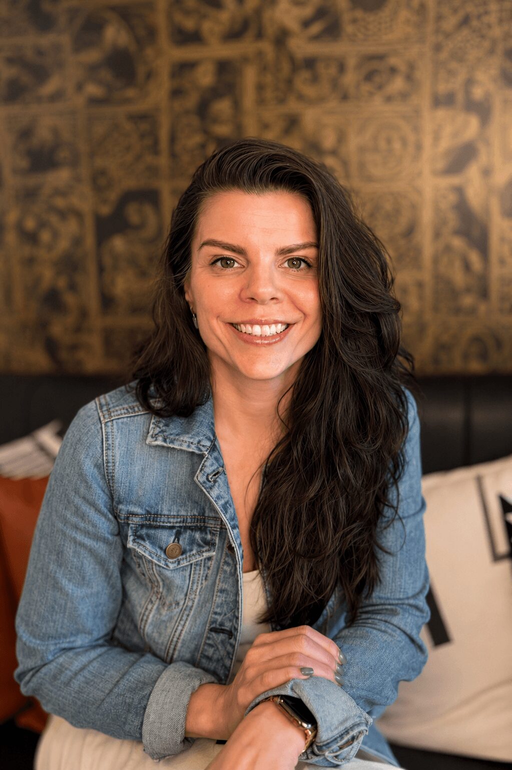 Smiling woman with long dark hair wearing a denim jacket, sitting indoors with a patterned wall behind her.