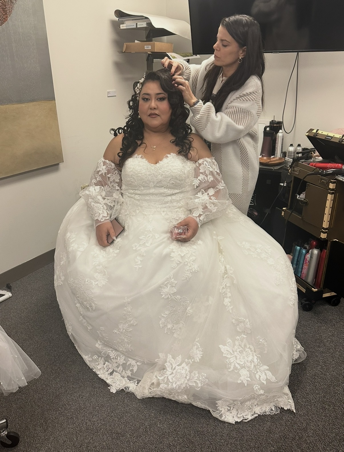 Woman in wedding dress getting her hair styled by a stylist in a room with a TV and shelves.
