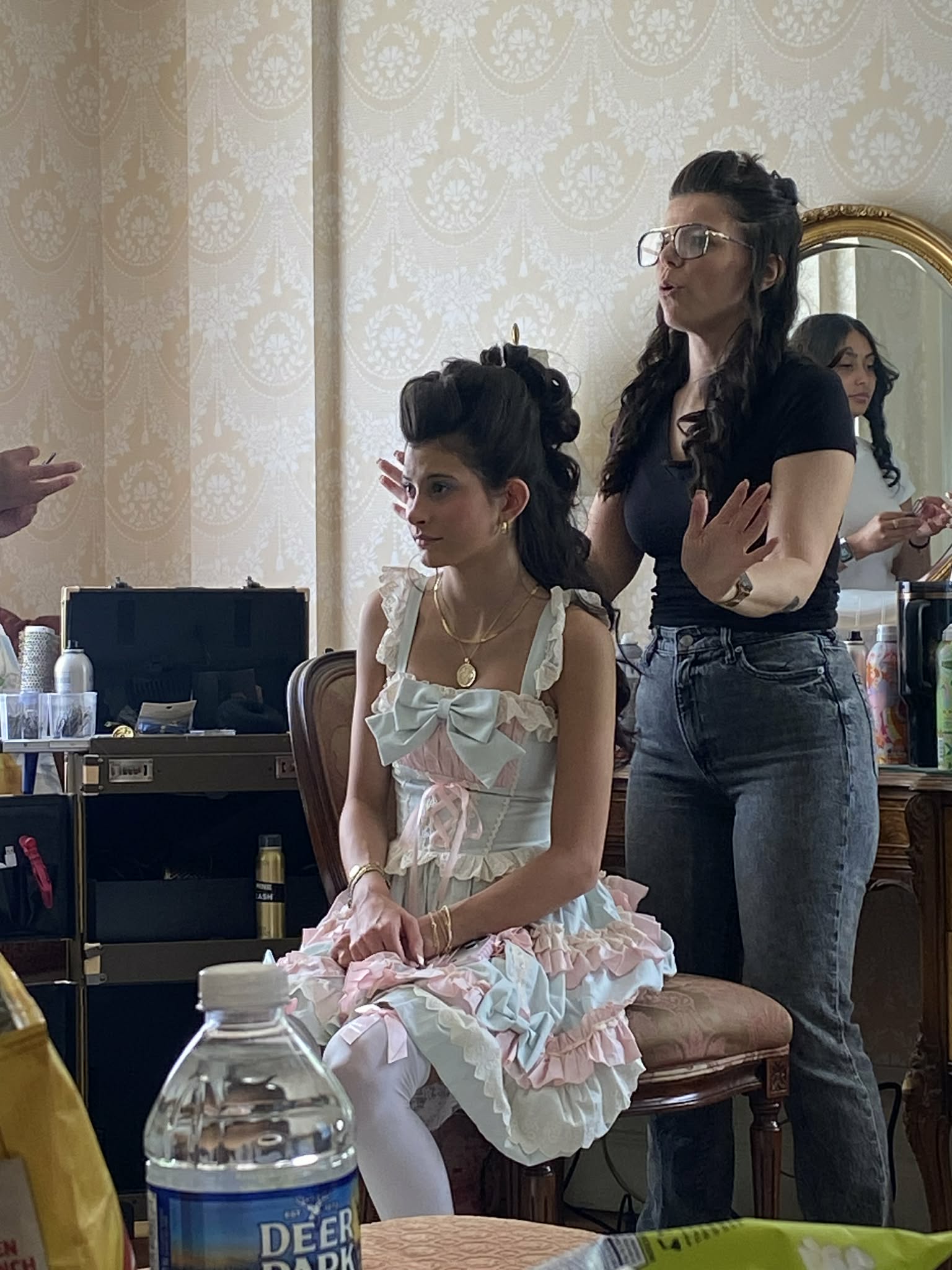 Two women in a room with a mirror, one sitting and one standing, surrounded by makeup and hair tools.