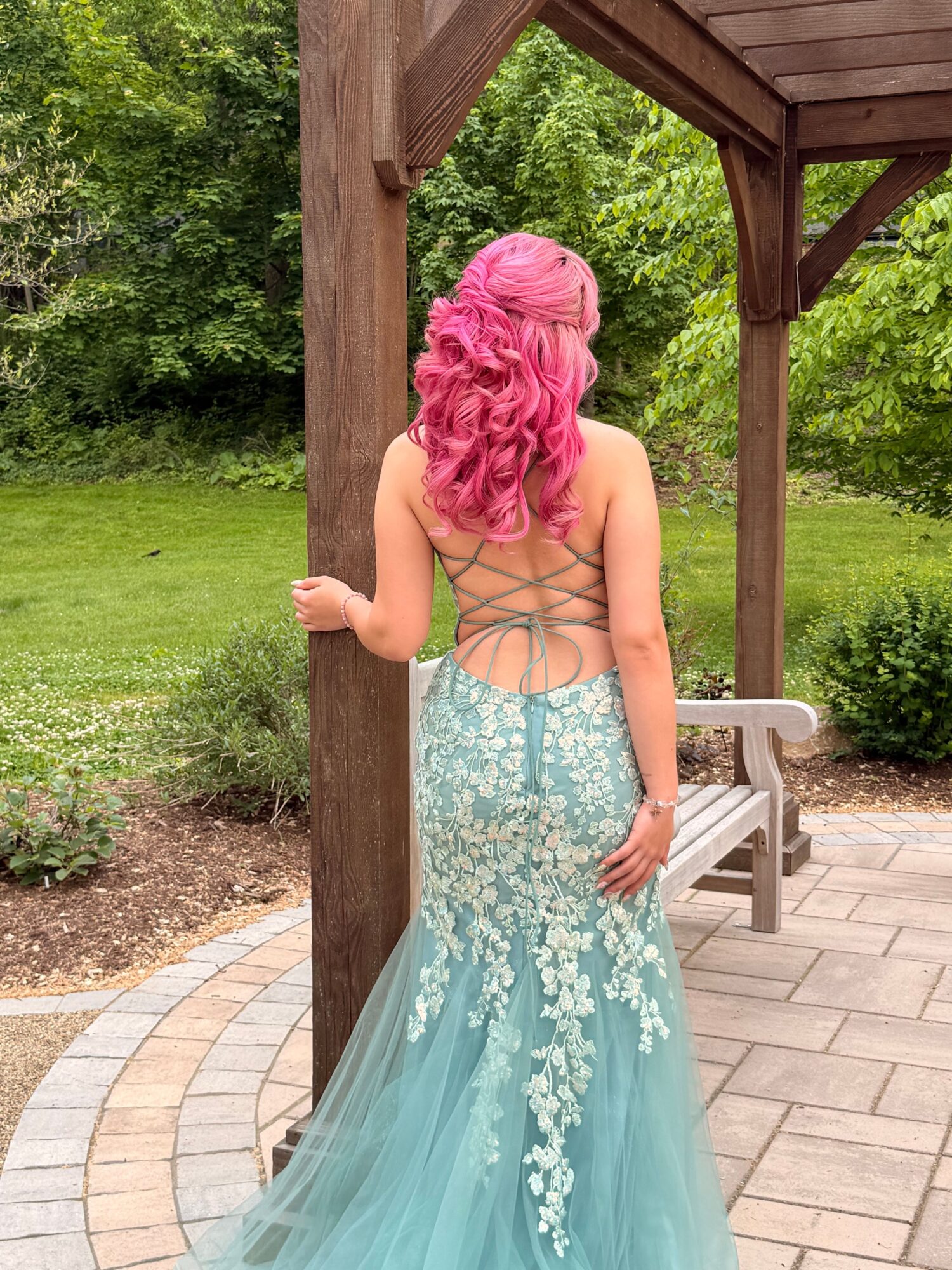 Woman with pink hair in a light blue dress standing outdoors near a wooden structure, greenery in background.
