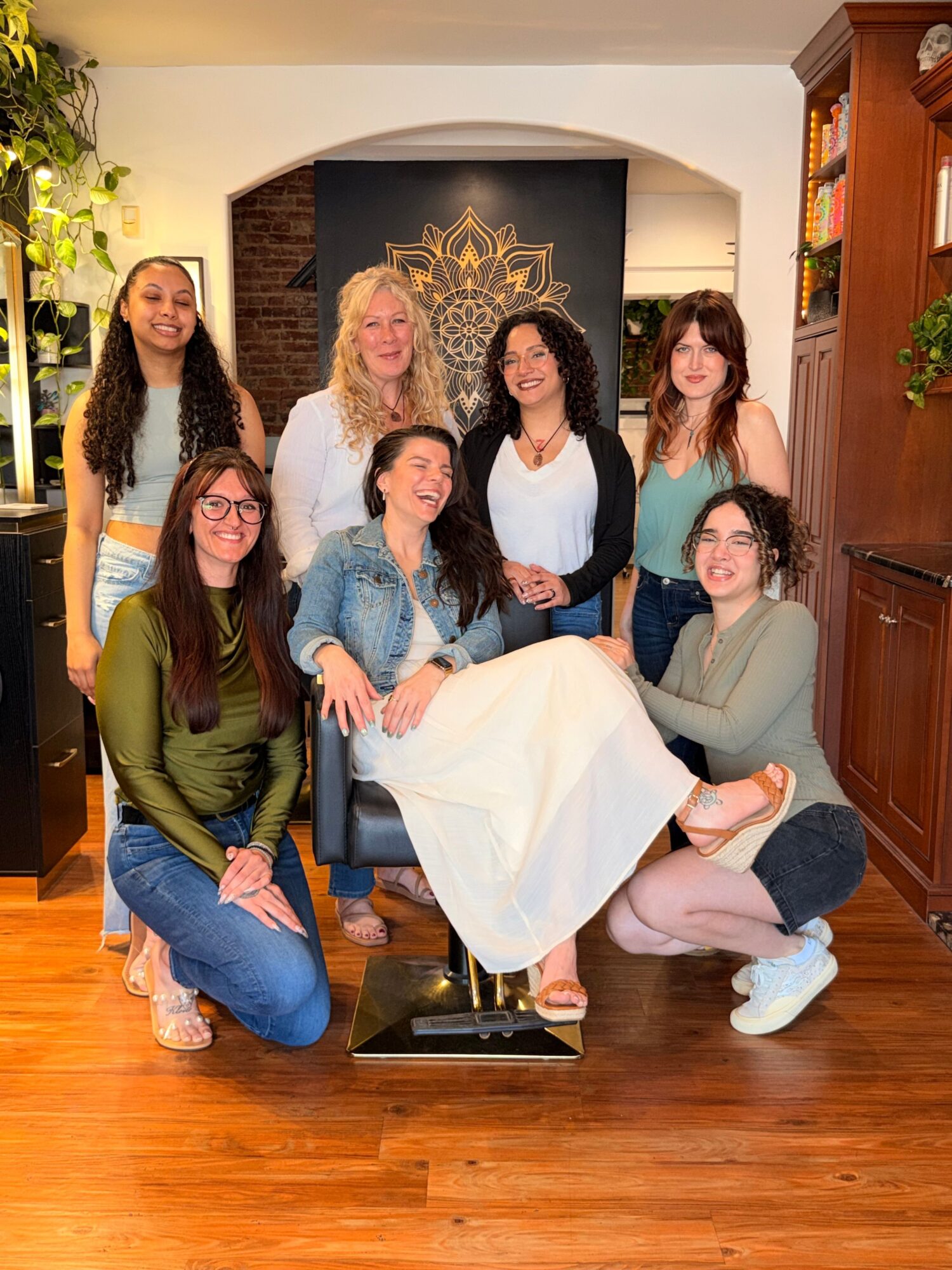 Group of six women sitting and standing around a woman seated in a white dress, in a room with wooden floors and a decorative wall.