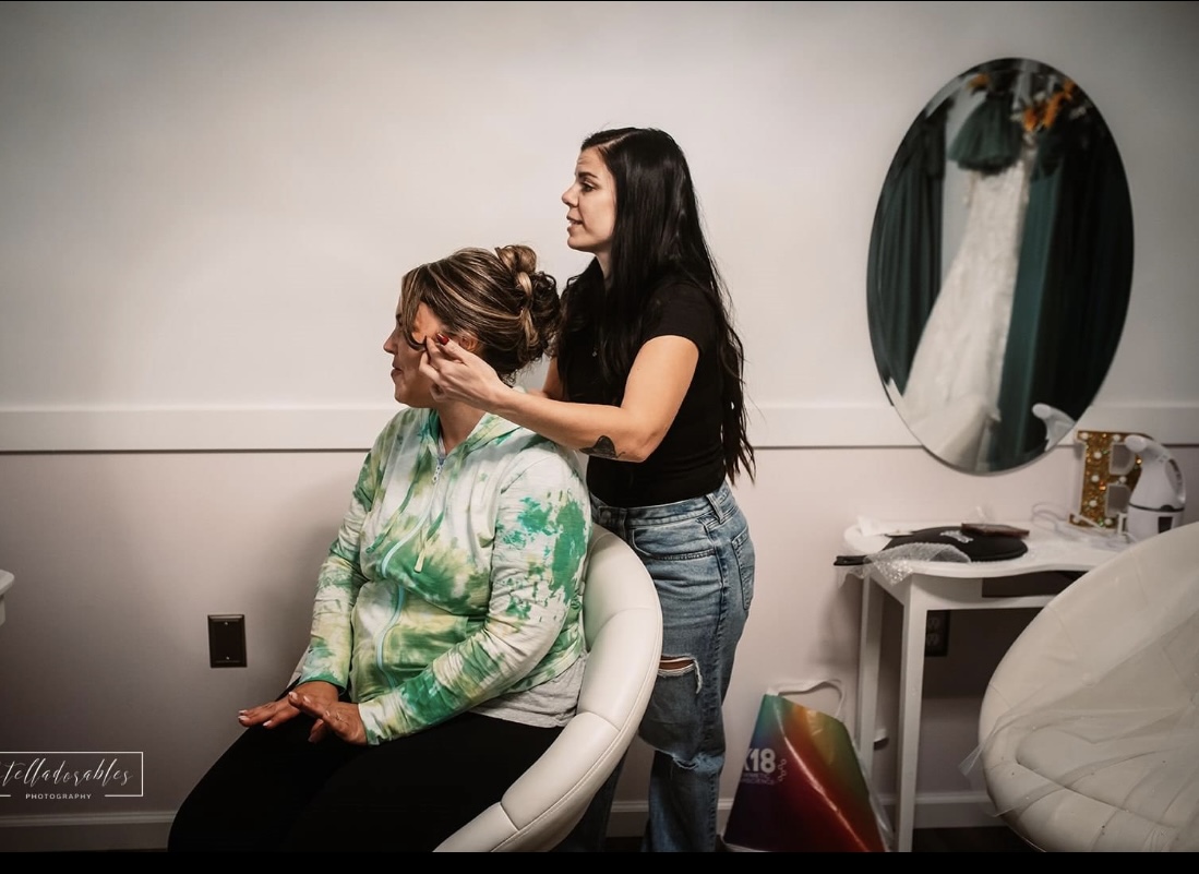 A woman is applying makeup to another woman seated in a chair, in a room with a mirror and various items on a table.